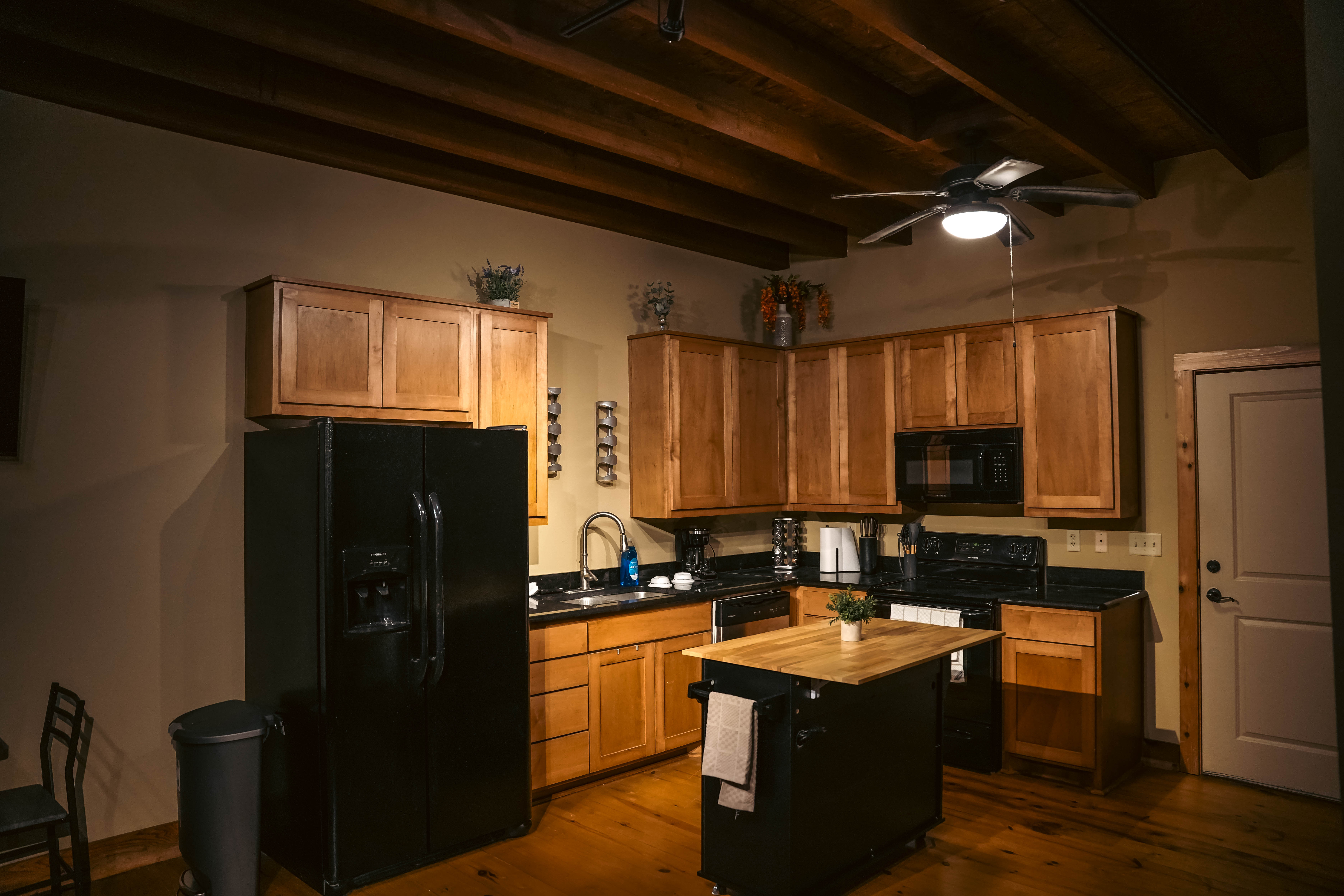 Cozy, rustic kitchen in an apartment featuring warm wood cabinets, a black refrigerator, dark countertops, and exposed wood ceiling beams.