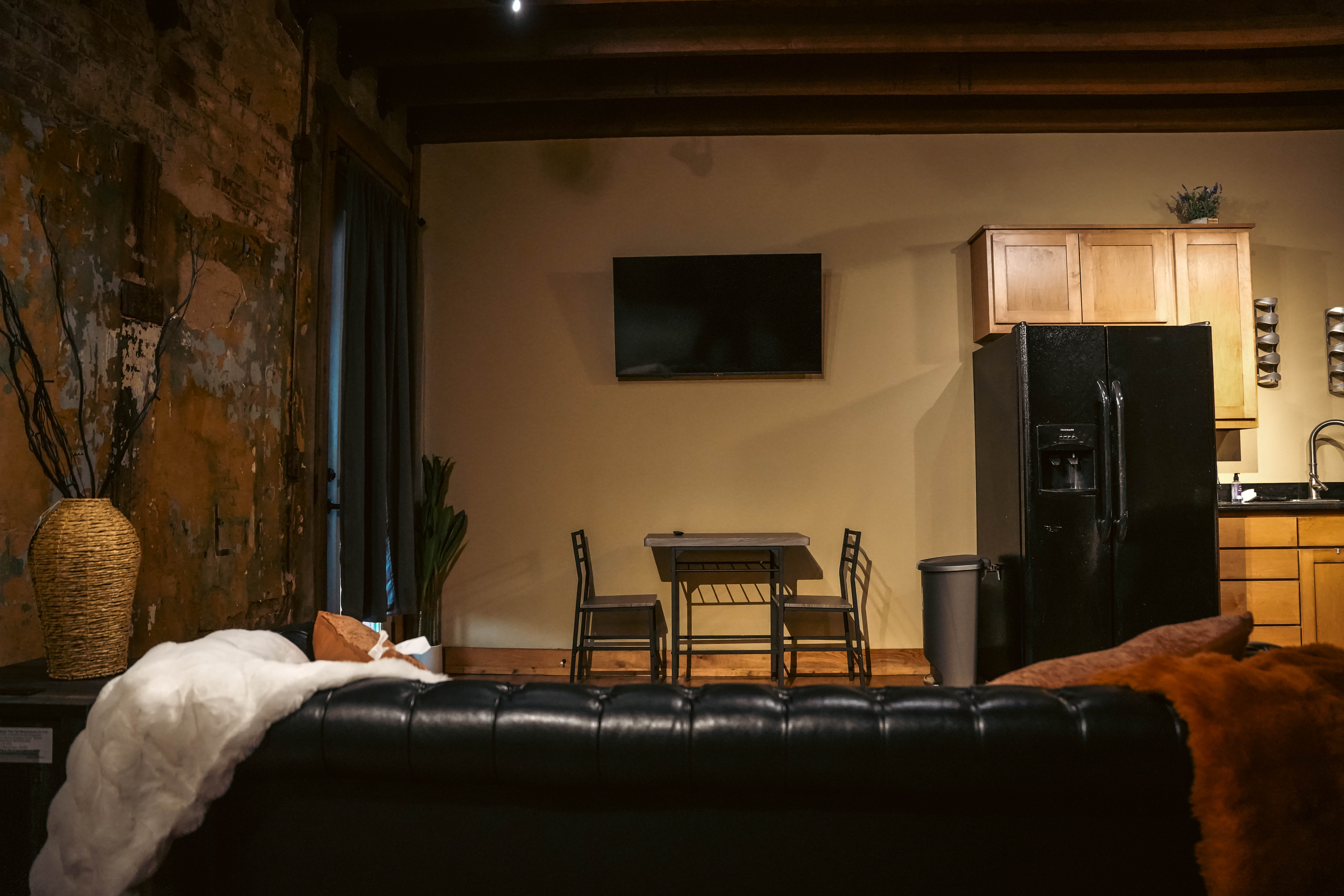Rustic, open-plan living area with a black leather sofa in the foreground, a small dining table and wall-mounted TV visible in the distance, and a stone wall texture.