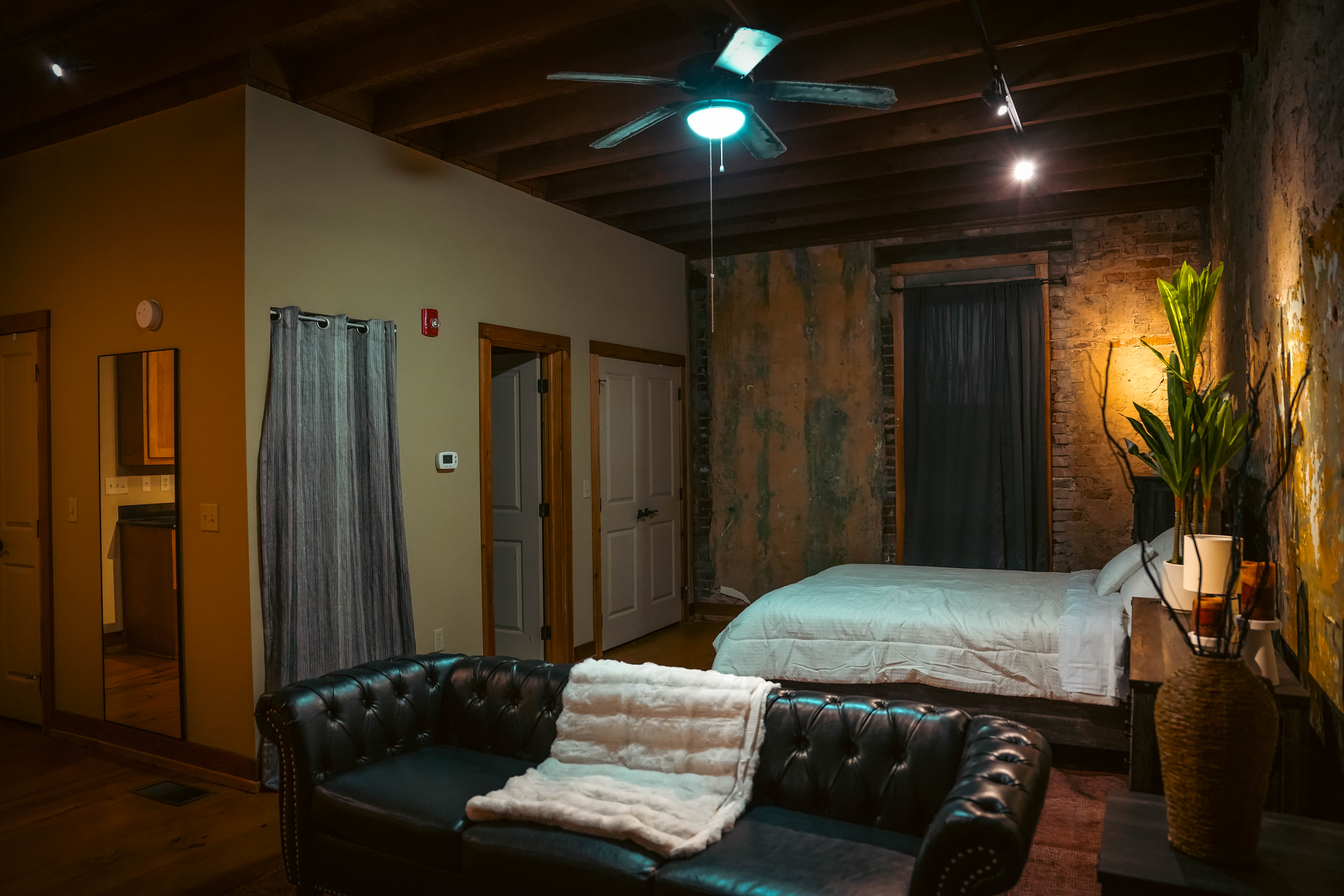 Cozy apartment bedroom with a black leather sofa, a white-sheeted bed, distressed walls, exposed wood ceiling beams, and a ceiling fan.