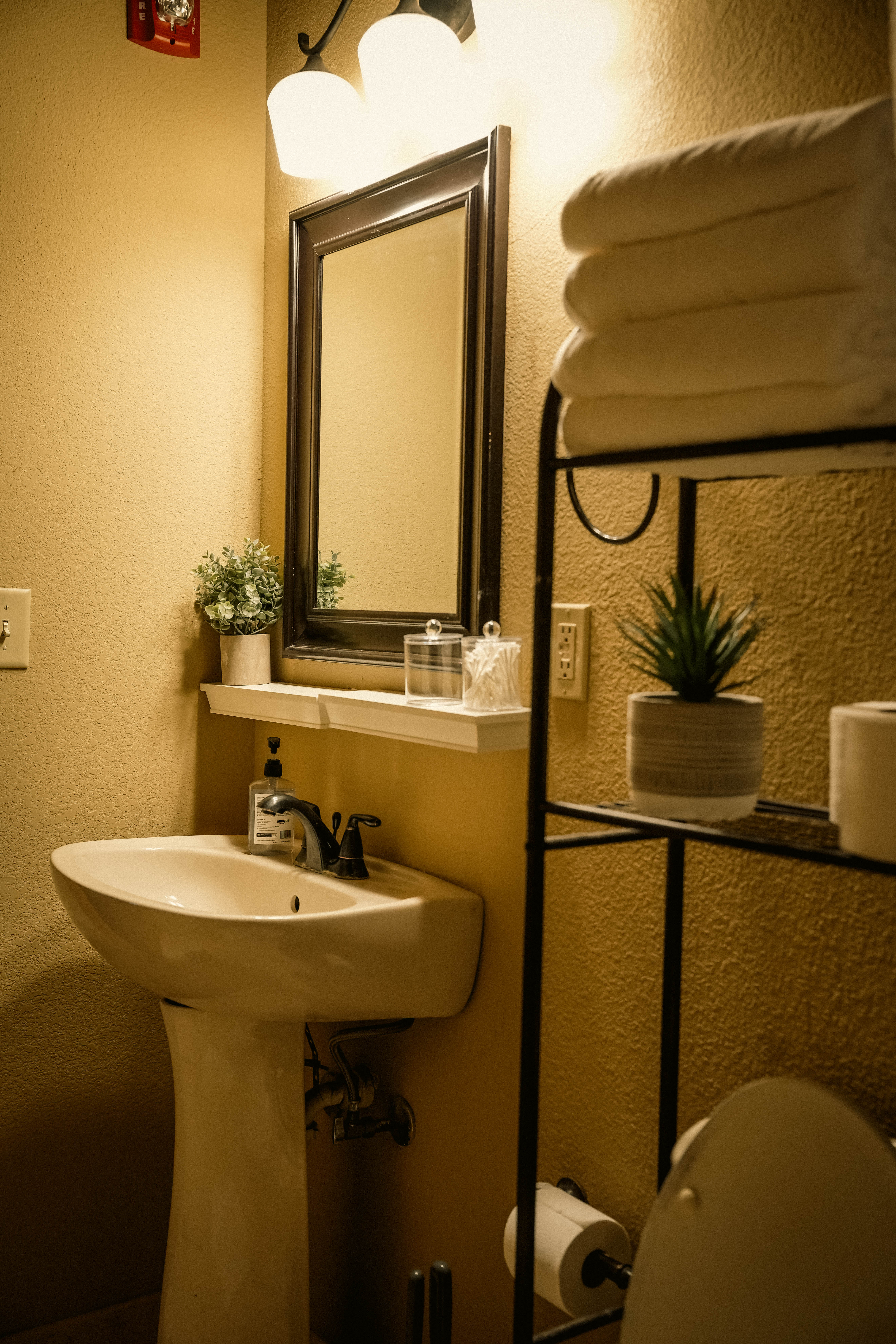 Warmly lit, small bathroom featuring a white pedestal sink, a framed mirror, a tiered black metal shelving unit with stacked towels, and small potted plants.