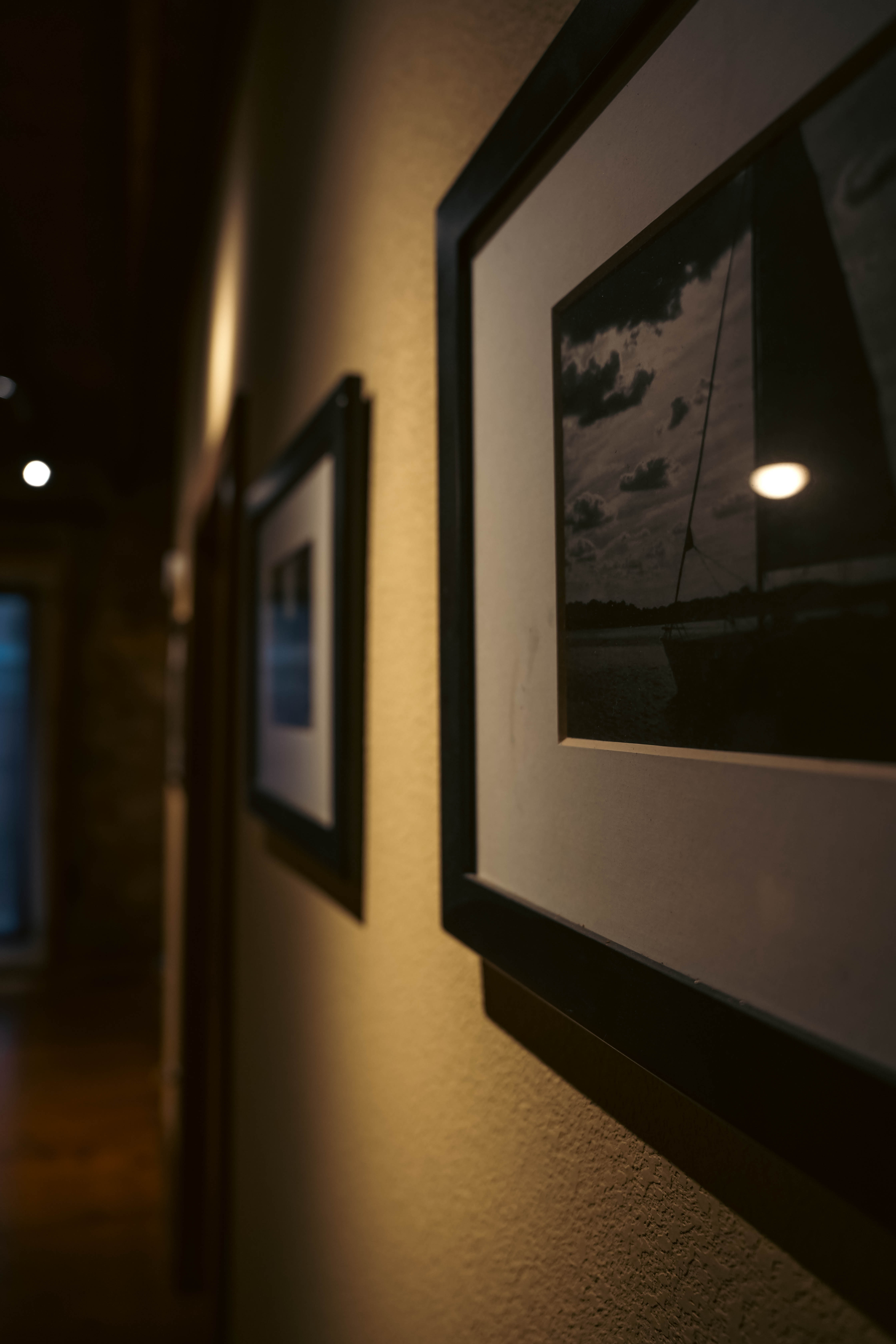 A dimly lit hallway with light-colored walls, featuring framed black and white photos hung along the side, leading to a wooden floor and a distant doorway.