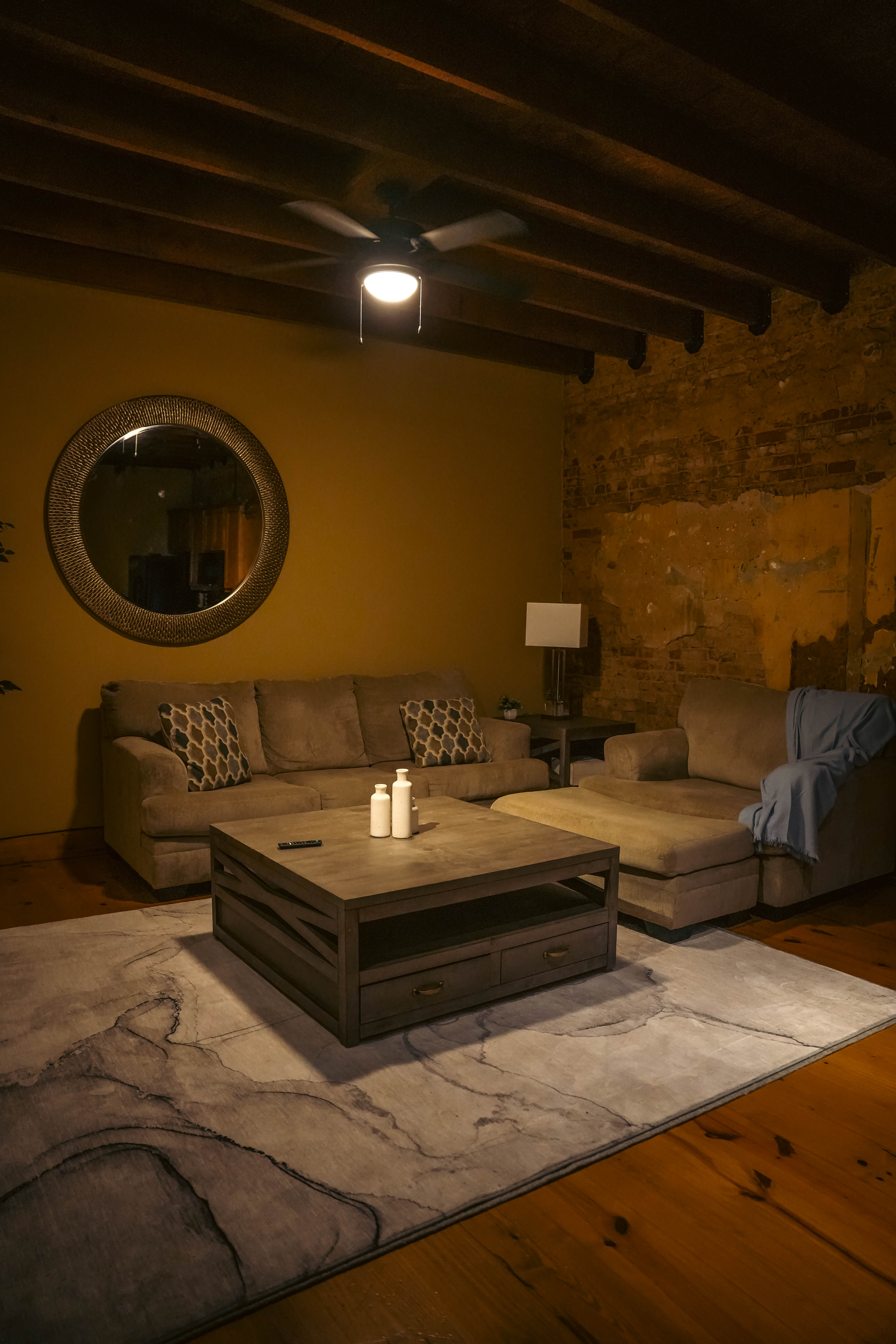Cozy, rustic living room featuring a tan sectional sofa, a dark wood coffee table with candles, a round mirror, exposed beams, and a stone accent wall.
