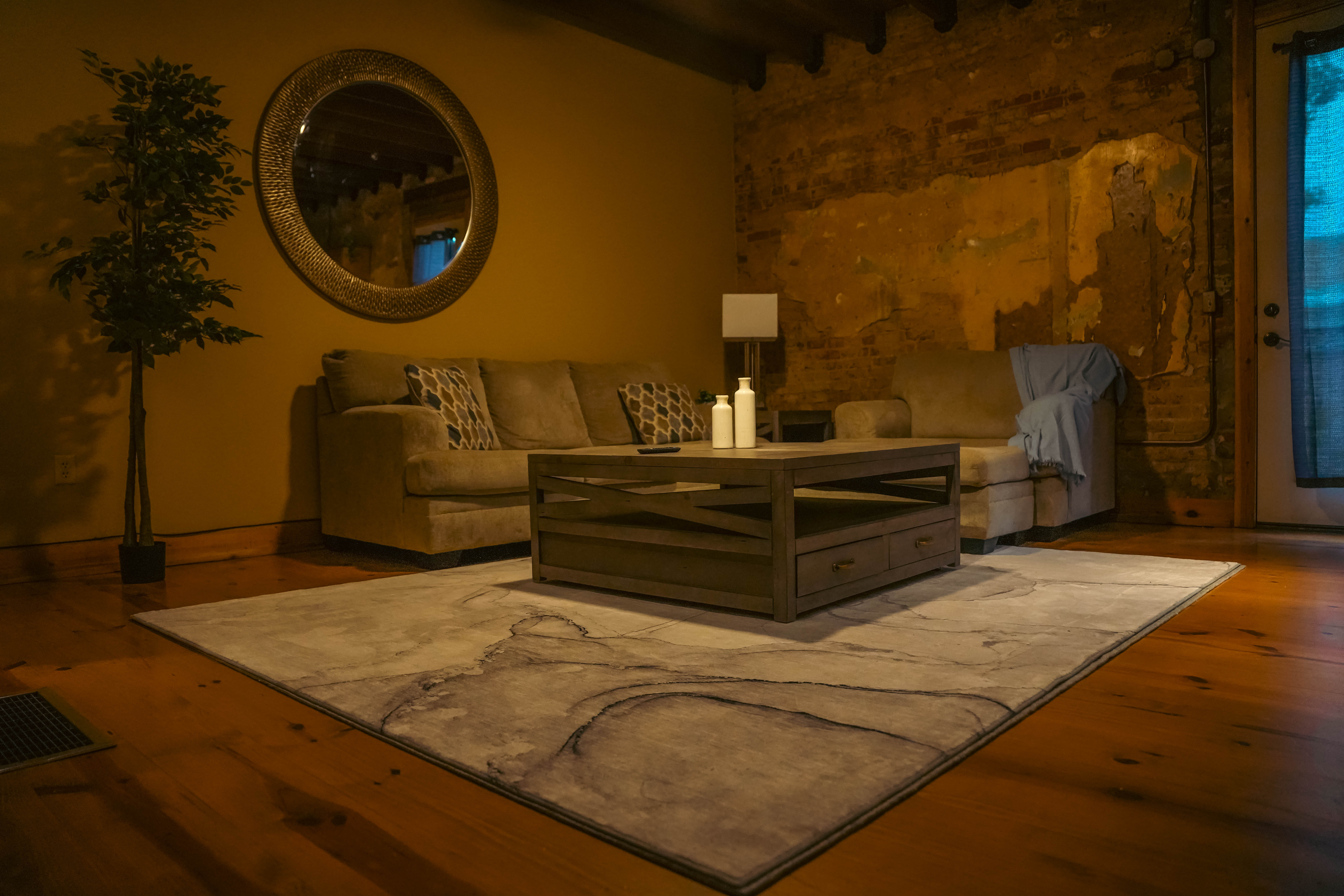 Rustic living space with a neutral sectional sofa, a distressed wood coffee table, a marble-pattern rug on a wooden floor, and a rough stone accent wall.