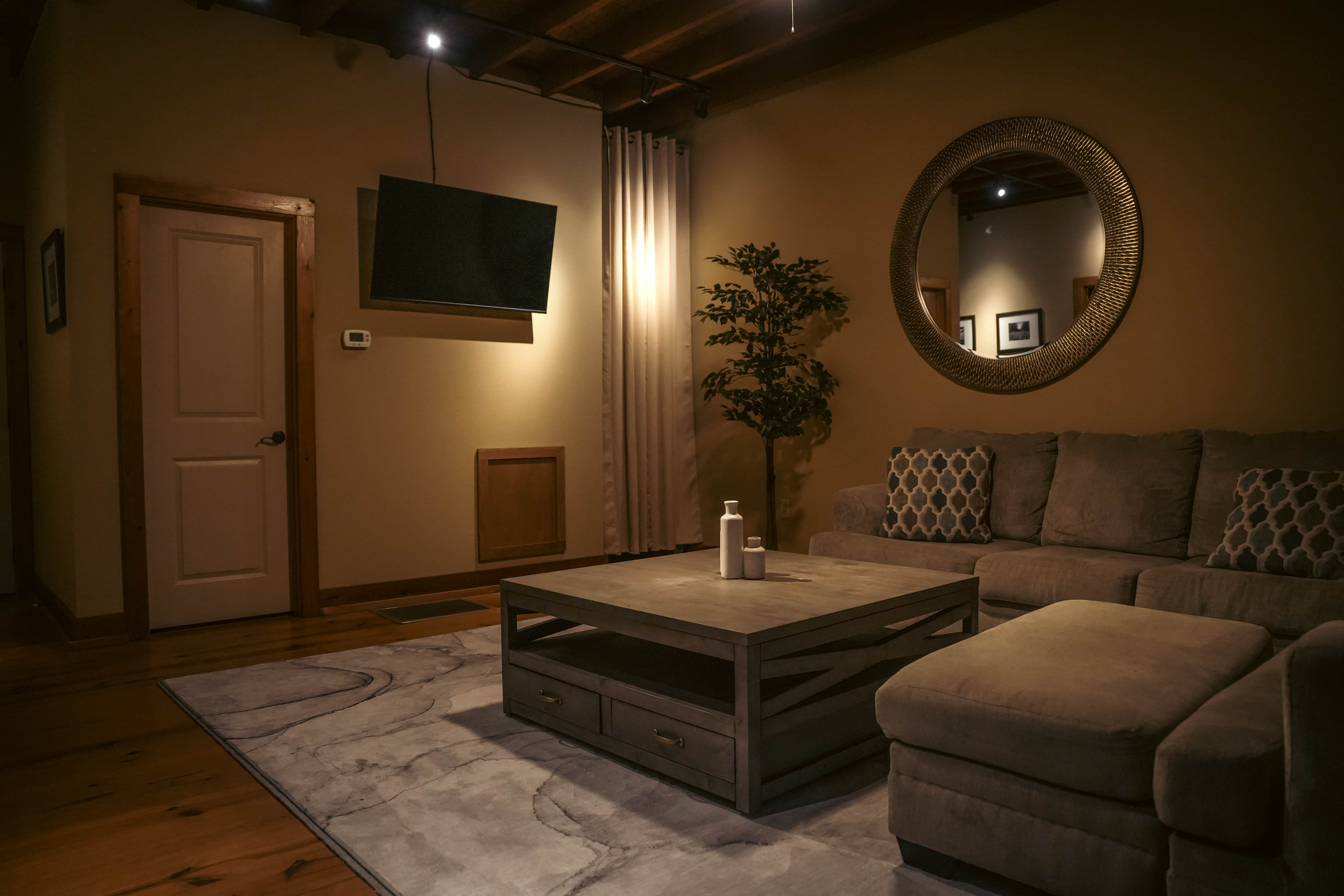 Cozy, dimly lit living room with a beige sectional, a rustic wooden coffee table, a large round mirror, a mounted TV, and wood floors.
