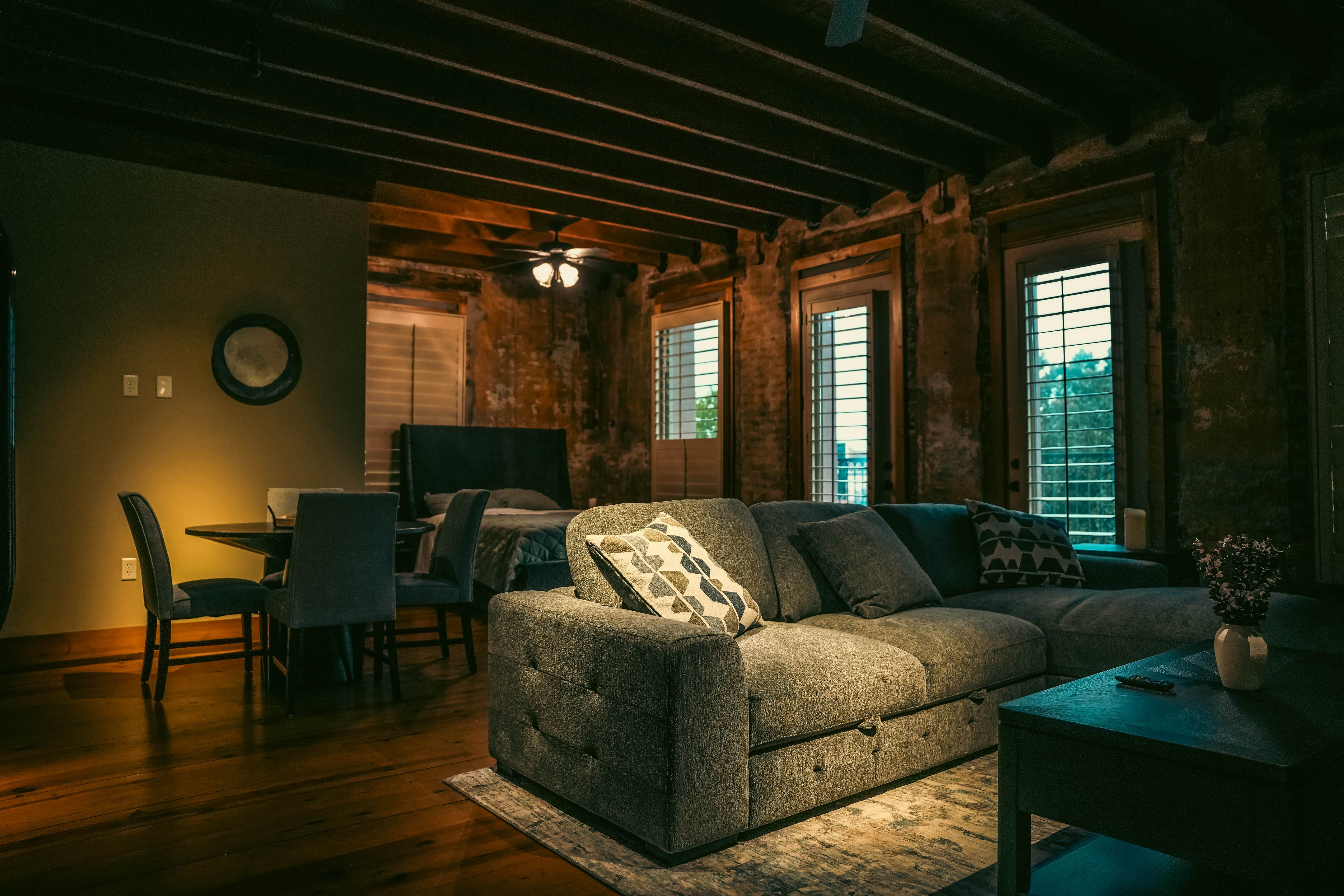 Rustic, industrial-style loft living space featuring a gray sectional sofa, a dining area behind it, exposed ceiling beams, and distressed brick walls with windows.