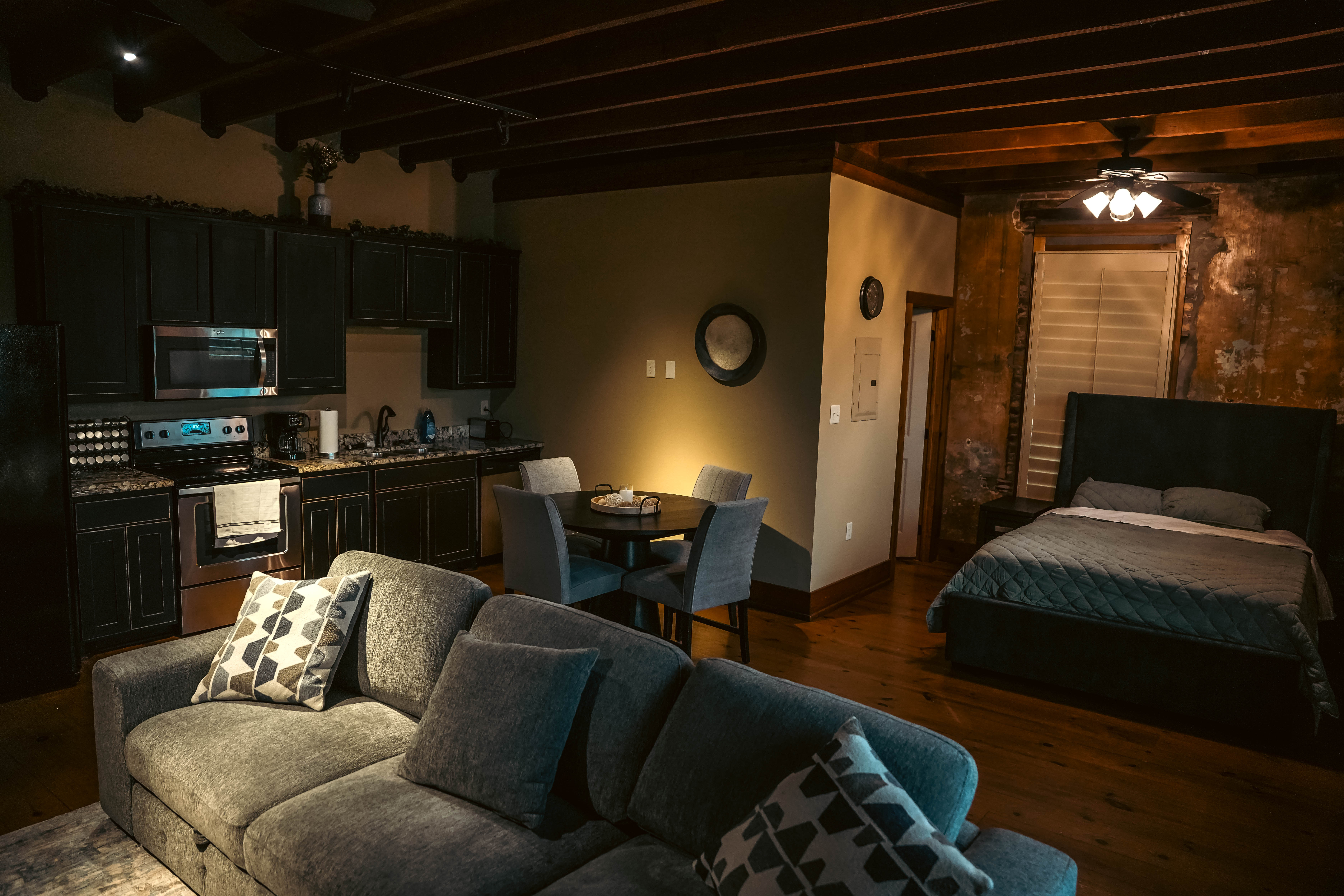 Open-concept studio apartment with a gray sofa in the foreground, a dark kitchen area, a small dining table, and a bed against a distressed brick wall.