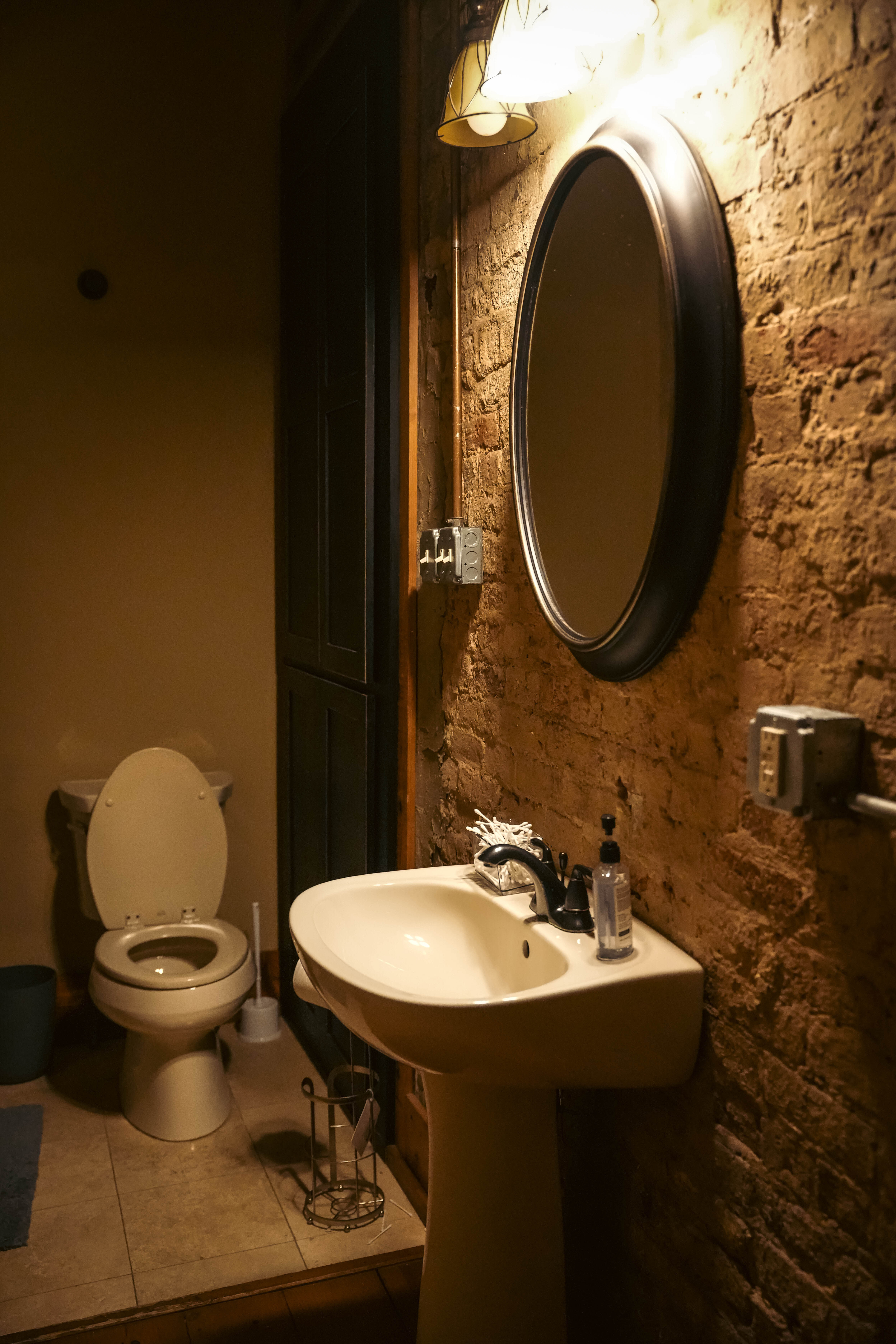 Rustic bathroom featuring a white pedestal sink, an oval mirror, a light fixture above it, and an exposed brick wall next to the toilet.