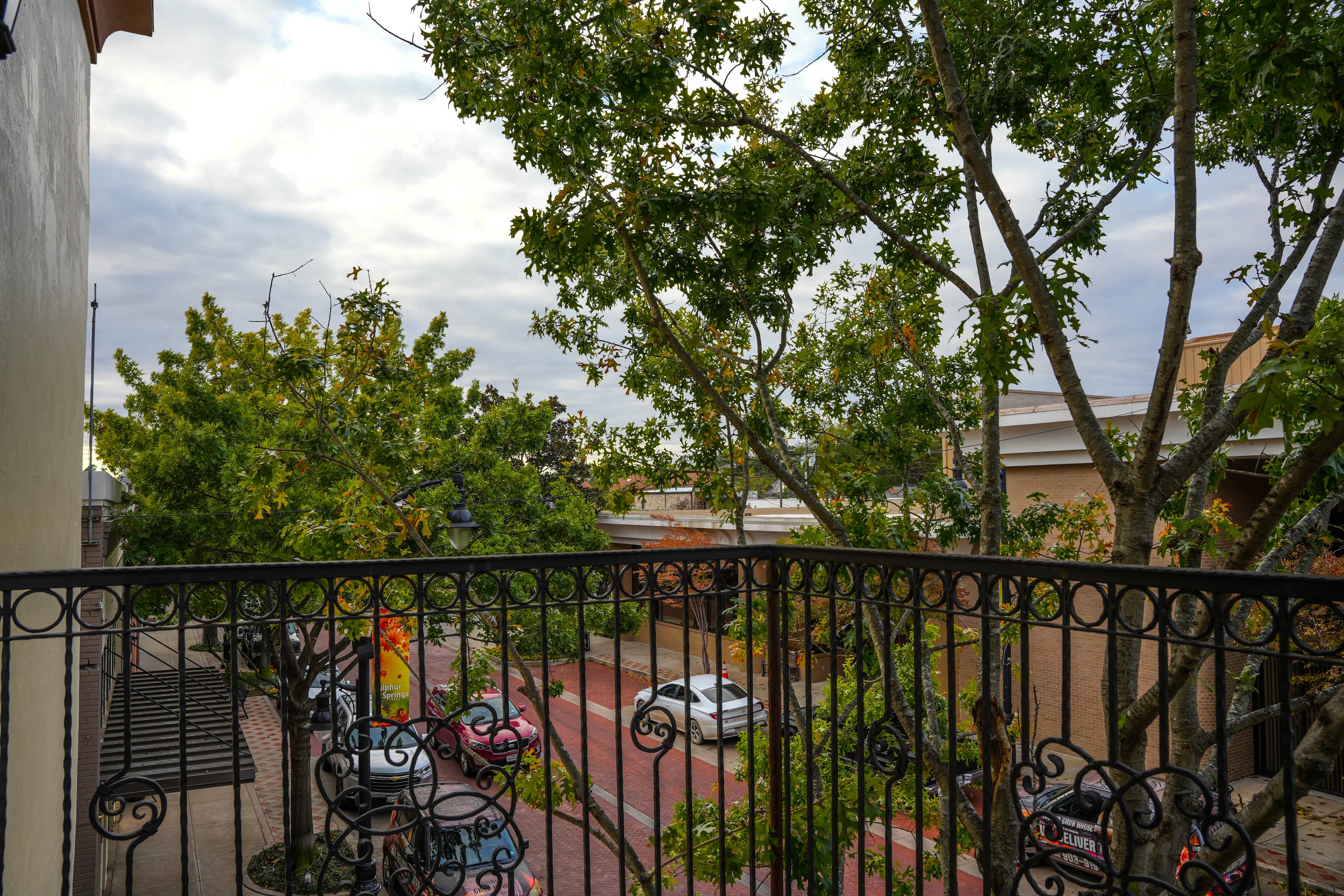 View from a balcony with a black wrought iron railing overlooking a tree-lined street with brick pavement and cars parked below on an overcast day.