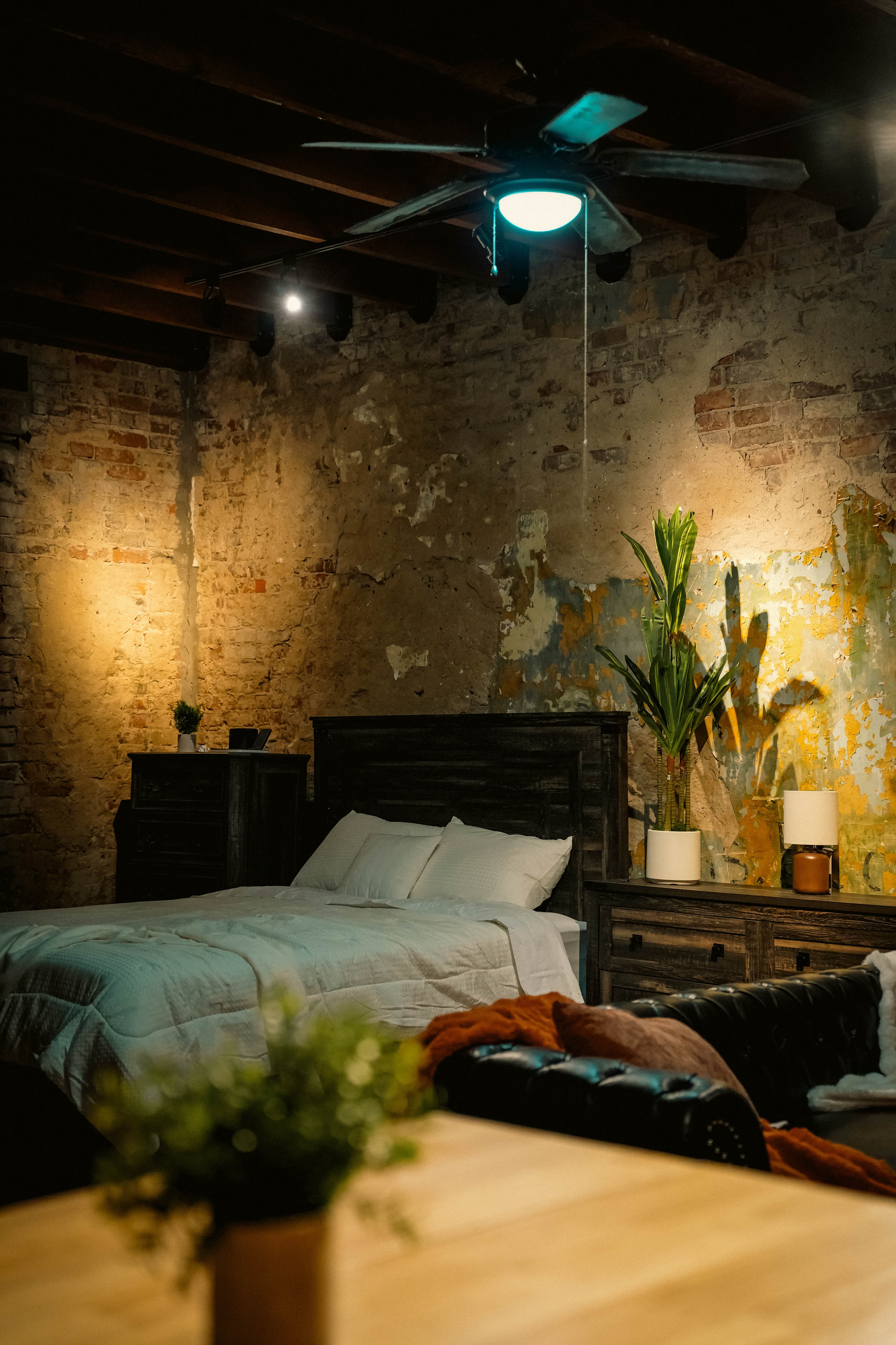 Rustic, dimly lit bedroom with distressed brick walls, a dark wood bed and nightstands, exposed ceiling beams, and a modern ceiling fan with a bright light.
