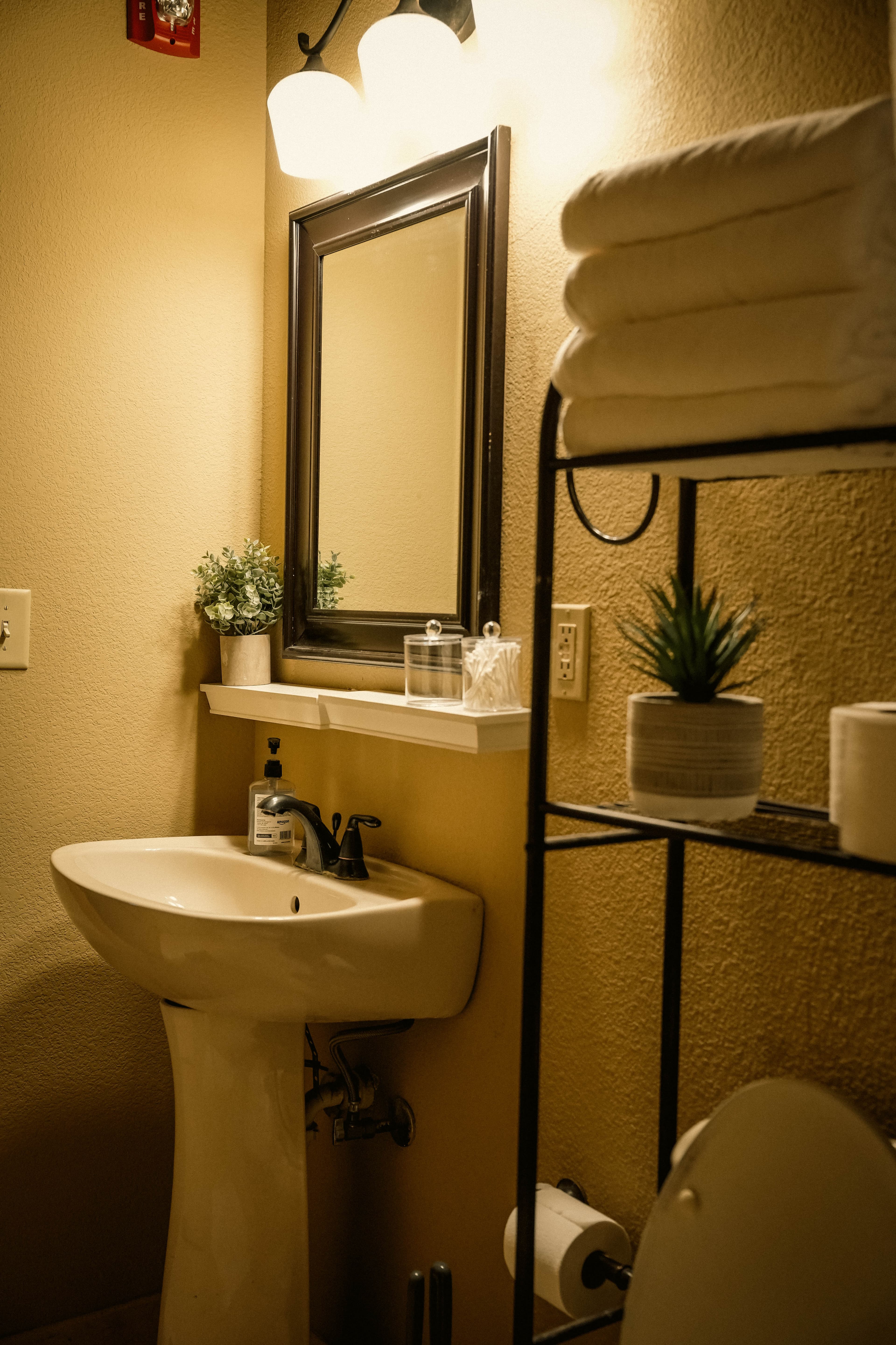 Warmly lit, small bathroom featuring a white pedestal sink, a framed mirror, a tiered black metal shelving unit with stacked towels, and small potted plants.