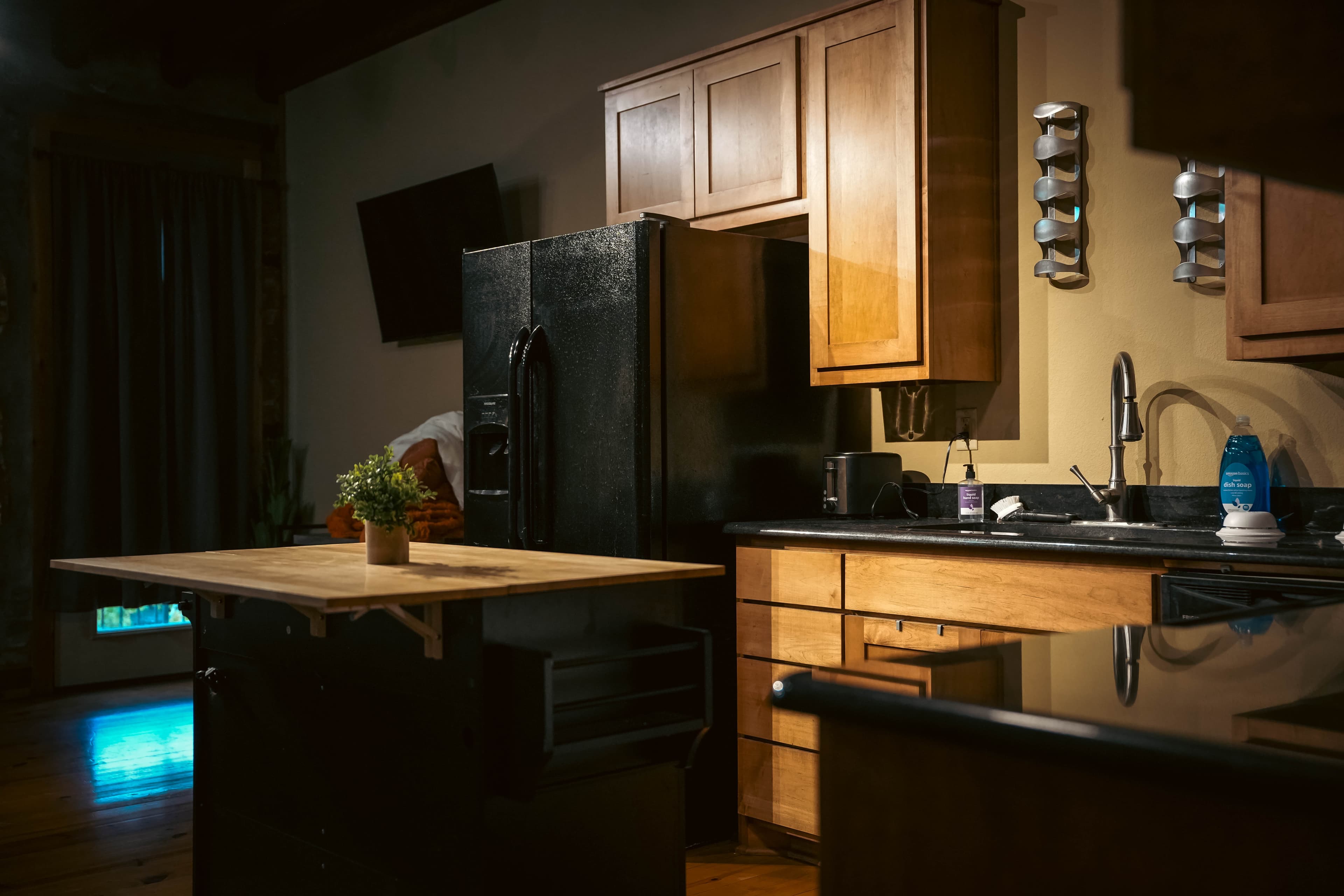 Dimly lit, cozy kitchen with warm wood cabinets, a black refrigerator, dark countertops, a wooden kitchen island, and a sink with a modern faucet.