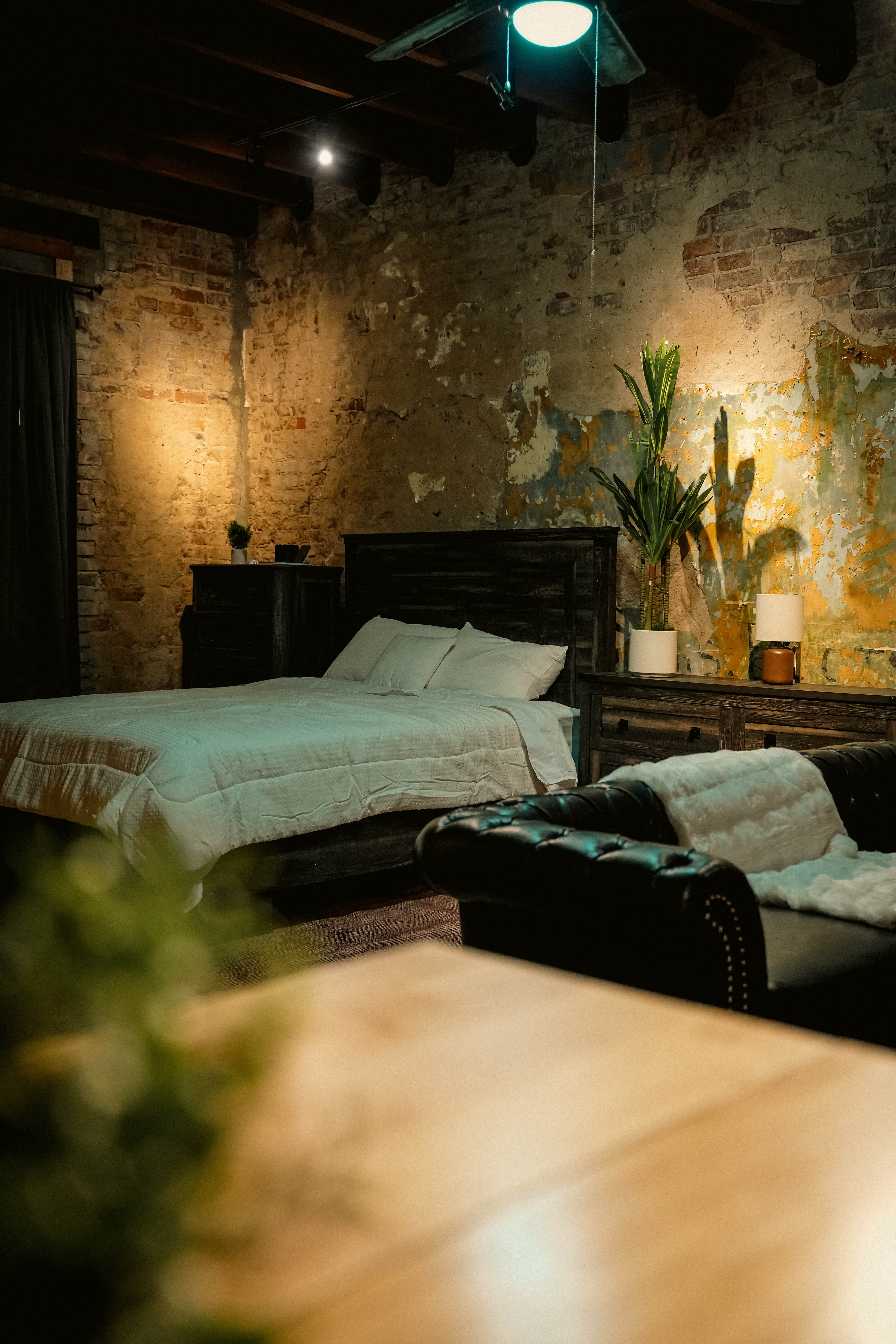 Dark, industrial-style bedroom featuring a bed with white bedding, a dark chaise lounge, distressed brick walls, and a large plant next to the bed.