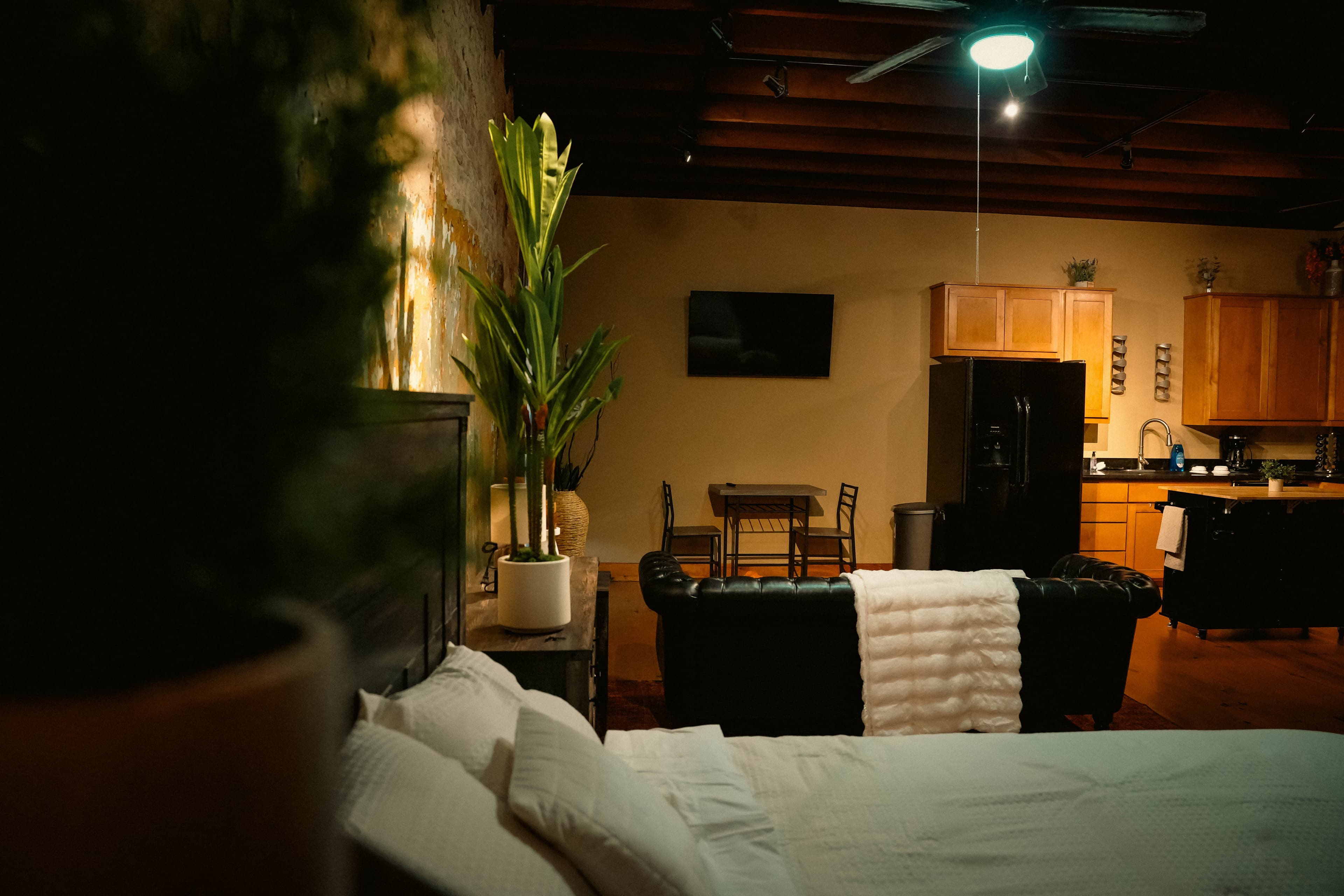 Open-plan apartment with a bedroom in the foreground, leading back to a living area with a dark sofa, a small dining set, and a full kitchen area.