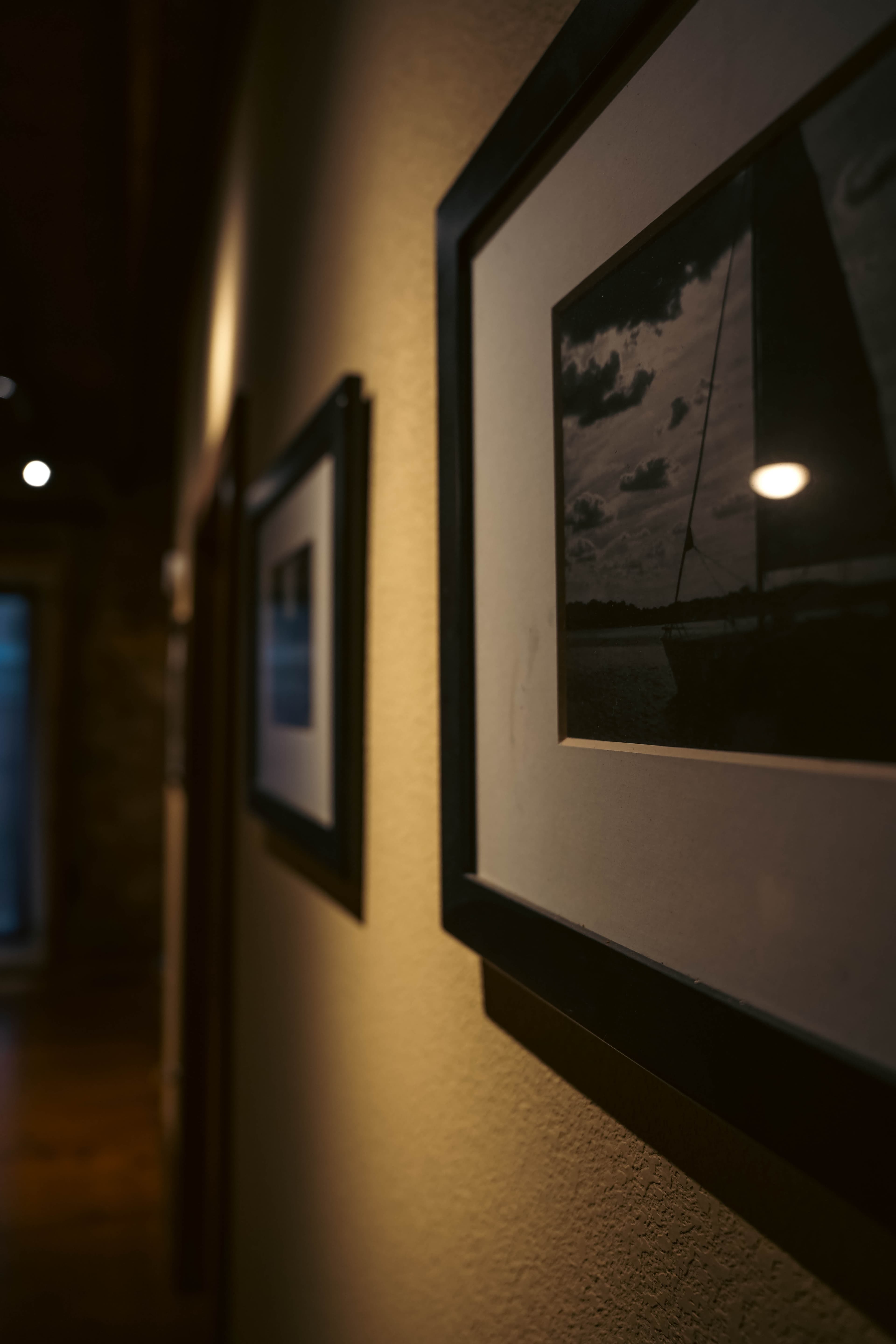 A dimly lit hallway with light-colored walls, featuring framed black and white photos hung along the side, leading to a wooden floor and a distant doorway.