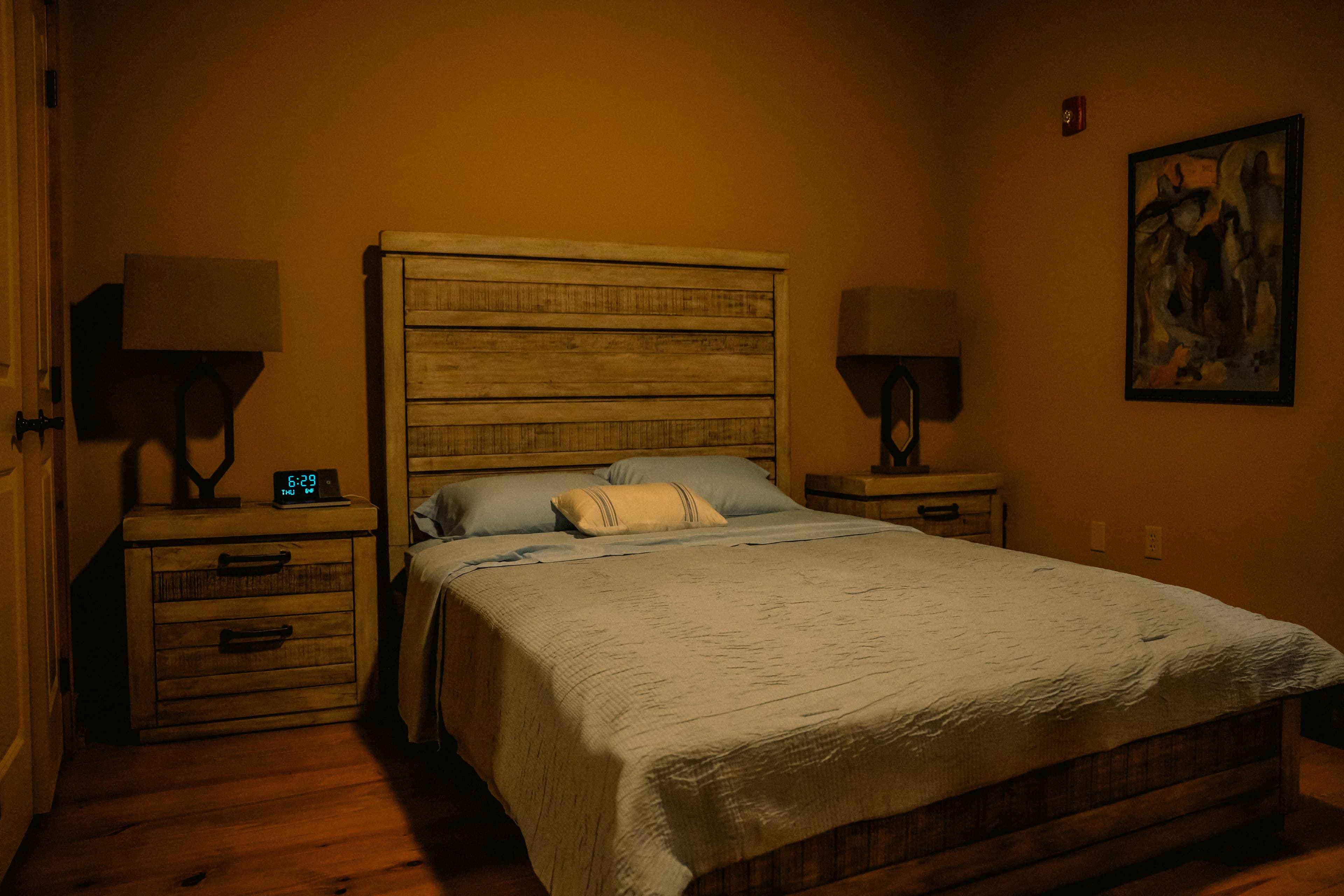 Cozy, dimly lit bedroom with a bed featuring a distressed wood headboard and frame, matching nightstands with lamps, and a piece of abstract art on the wall.