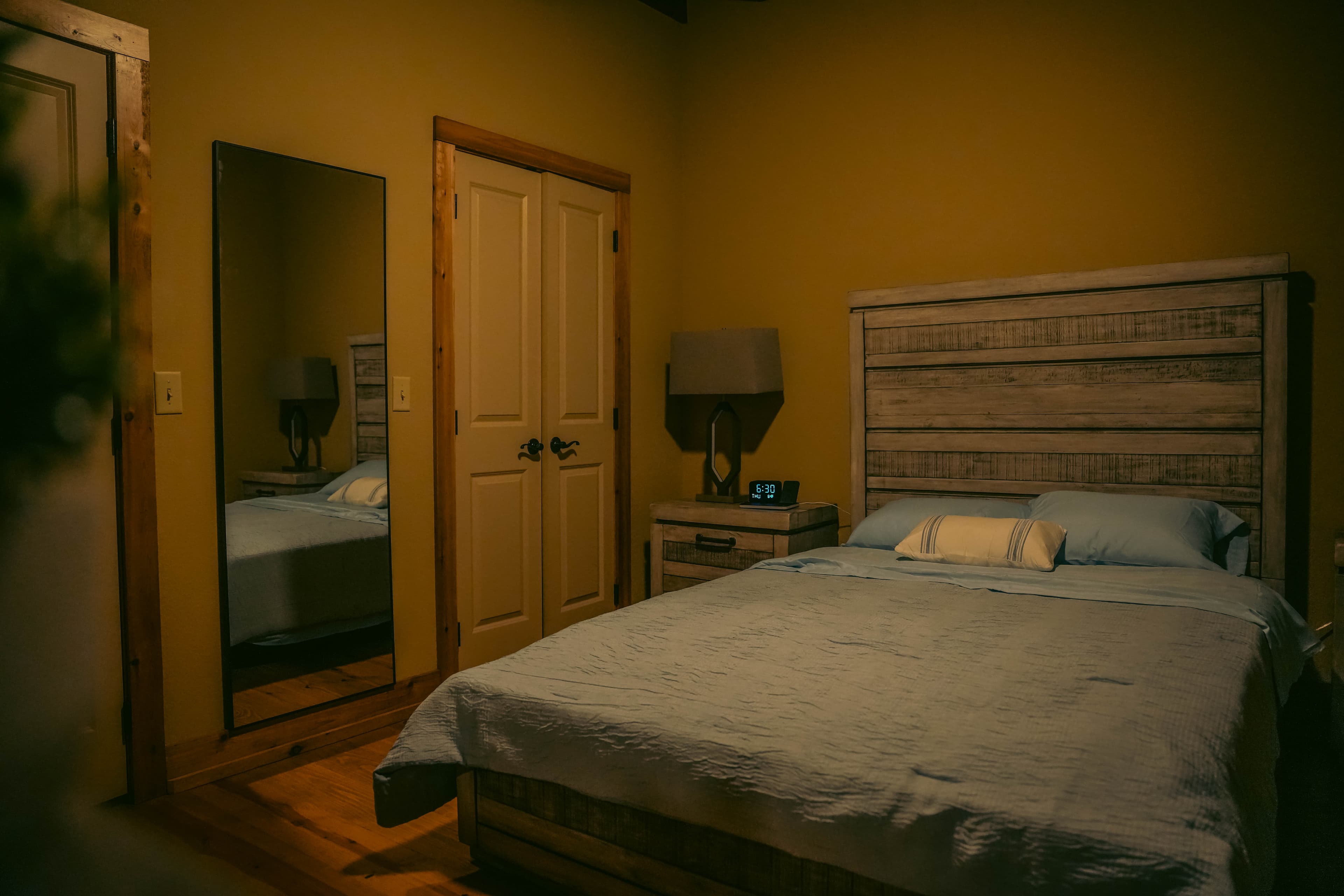 Rustic bedroom with a distressed wood bed frame and light blue bedding, a full-length mirror, closed double closet doors, and wood flooring.