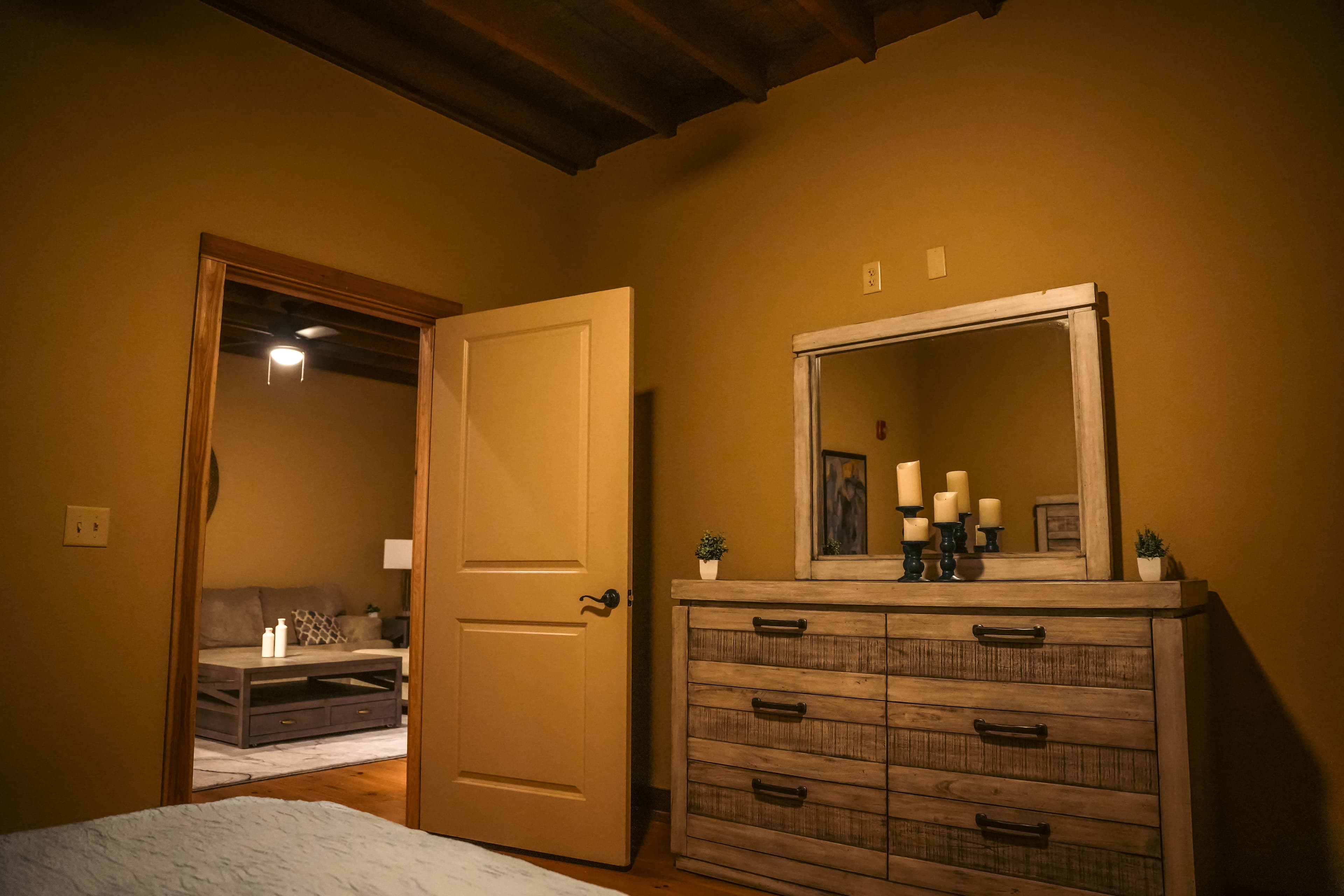 Bedroom perspective showing an open door leading into a living room, with a rustic, distressed wood dresser, mirror, and candles in the foreground.