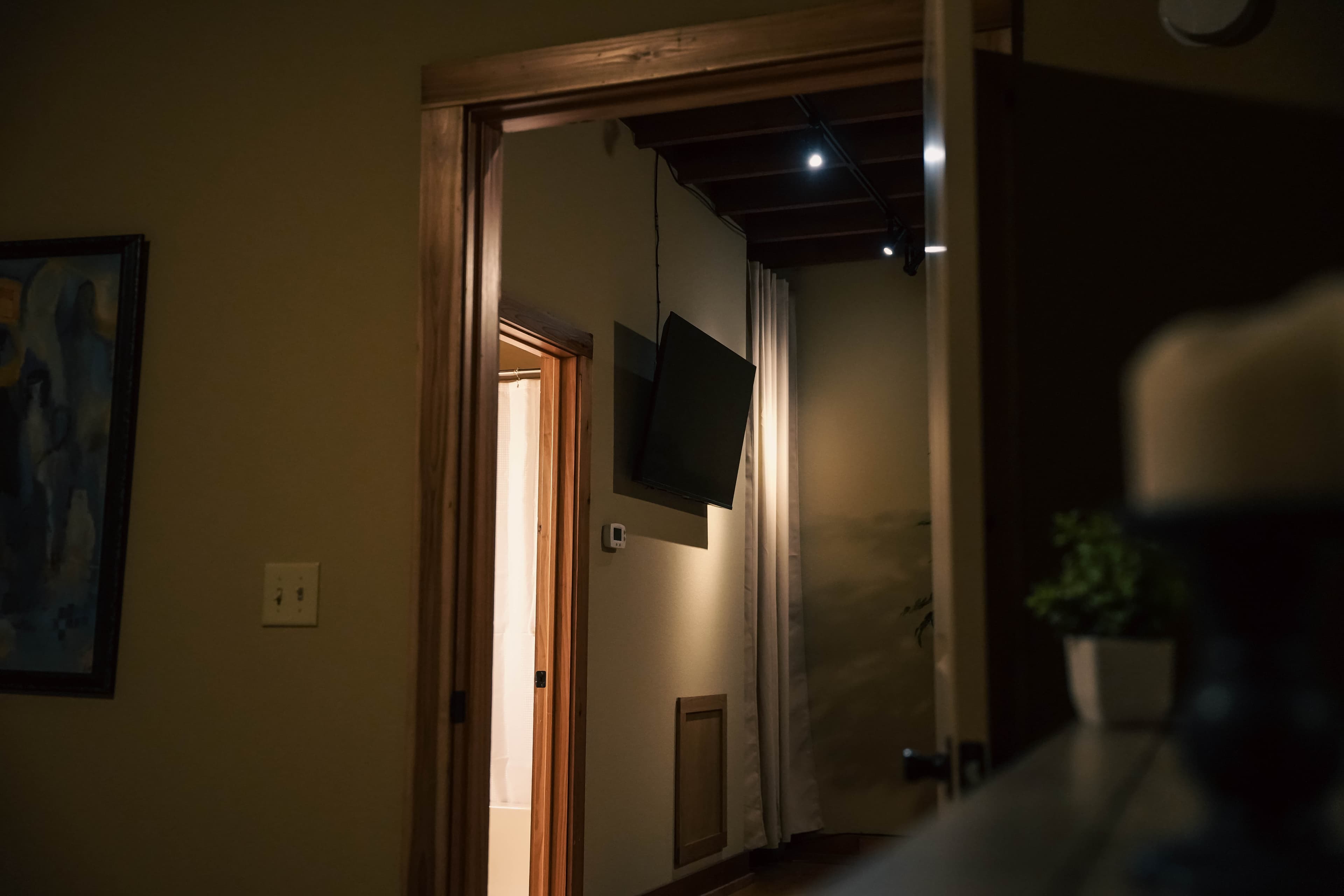 Interior shot looking through a doorway into a hallway with dark wood trim, mounted TVs, distressed walls, and exposed ceiling beams in the background.