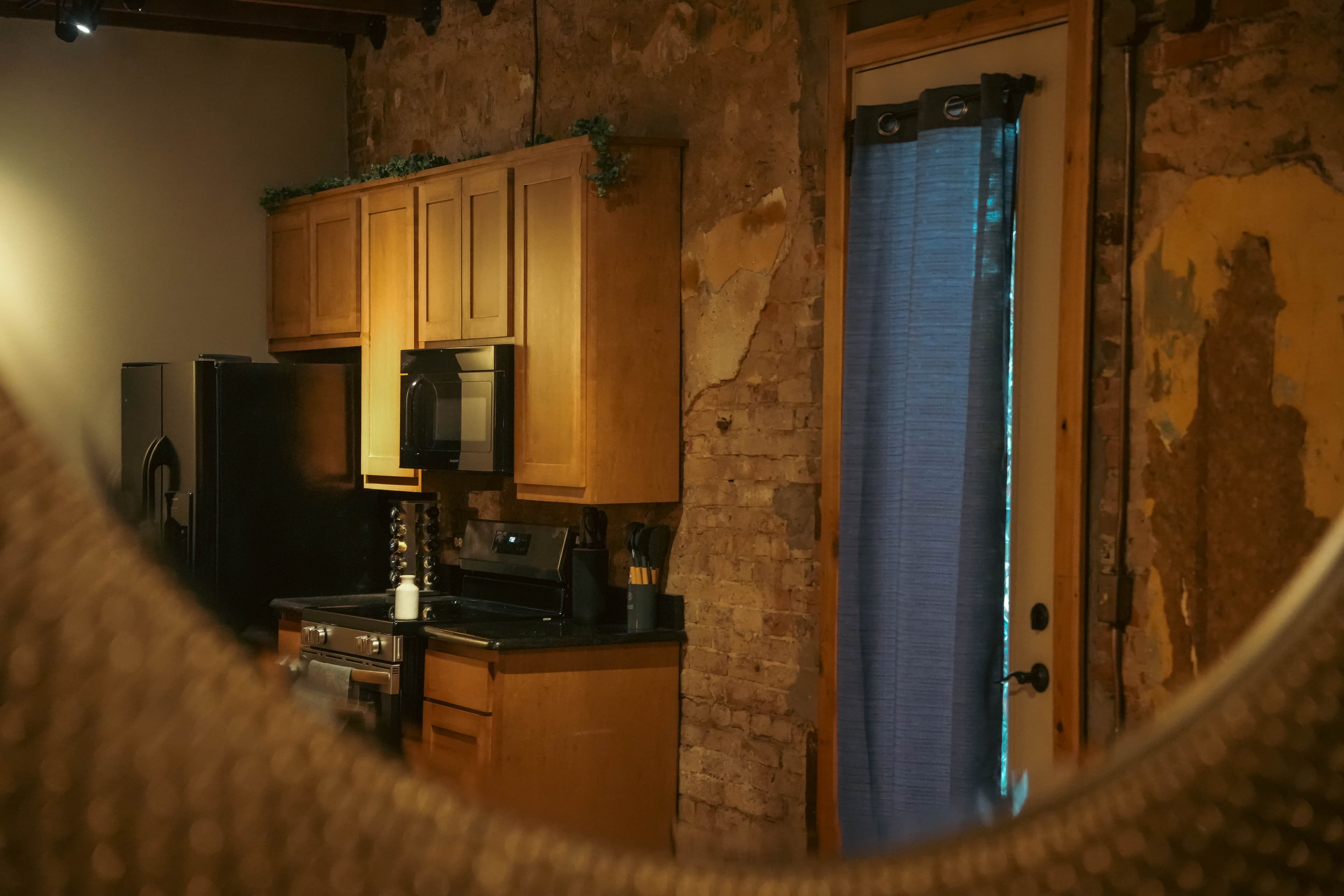 View into a rustic kitchen with light wood cabinets, black appliances, and a distressed brick wall, seen through a blurred mirror.