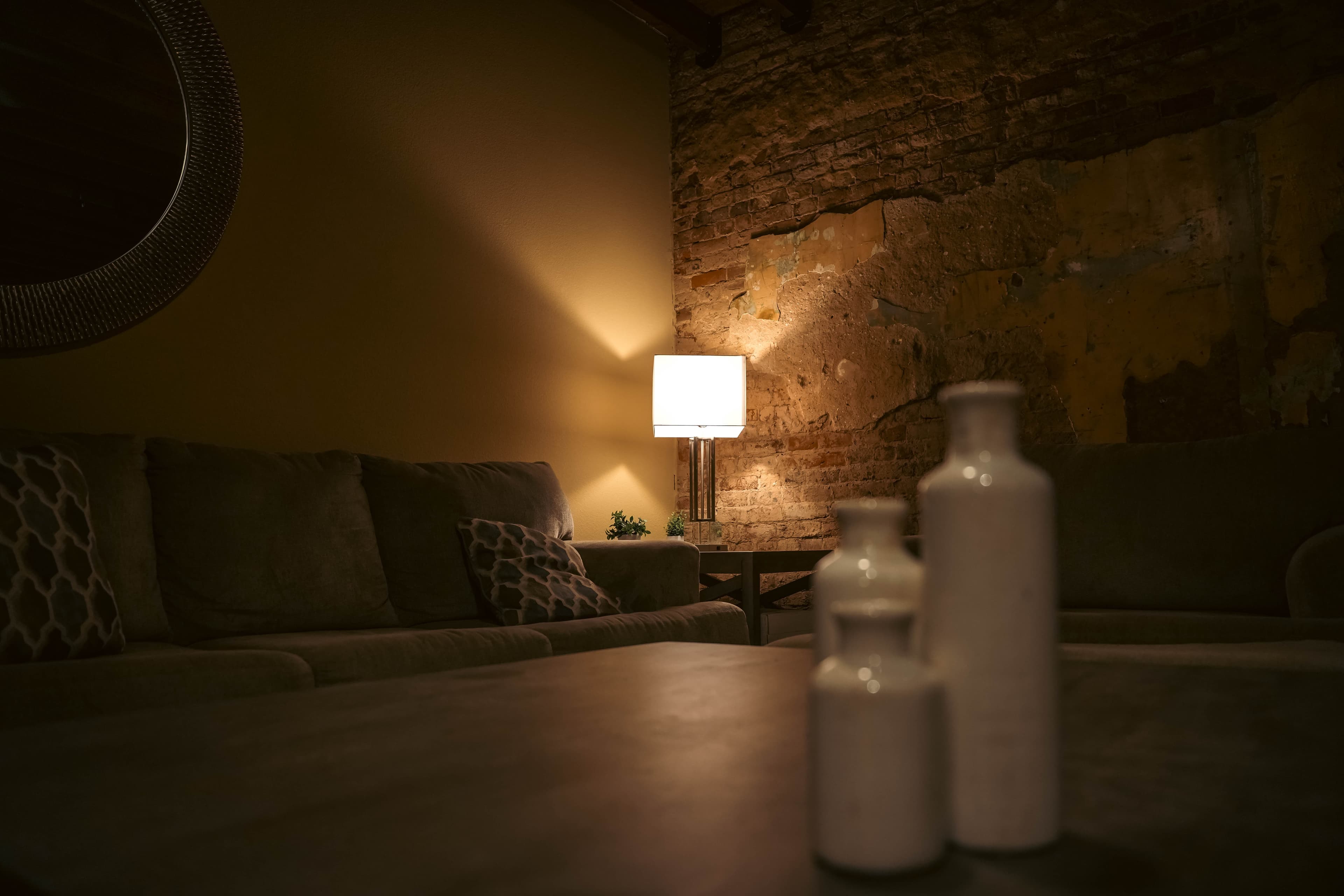 Cozy, dimly lit living room with white decorative vases in the foreground, a corner lamp illuminating a sectional sofa, and a distressed brick accent wall.