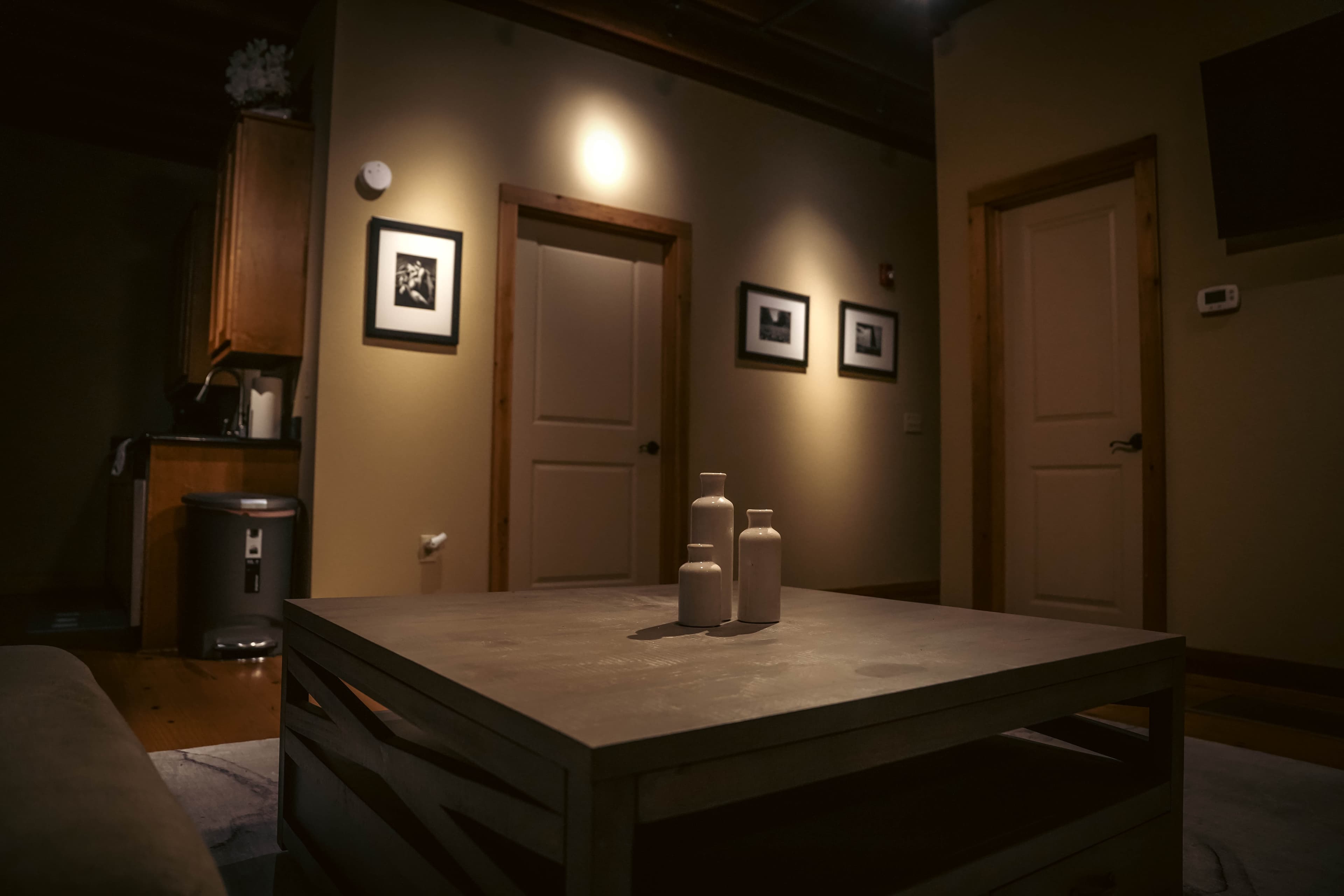 Dimly lit interior shot focusing on a square wooden coffee table with decorative vases, framed black and white photos on the wall, and two closed light-colored doors.