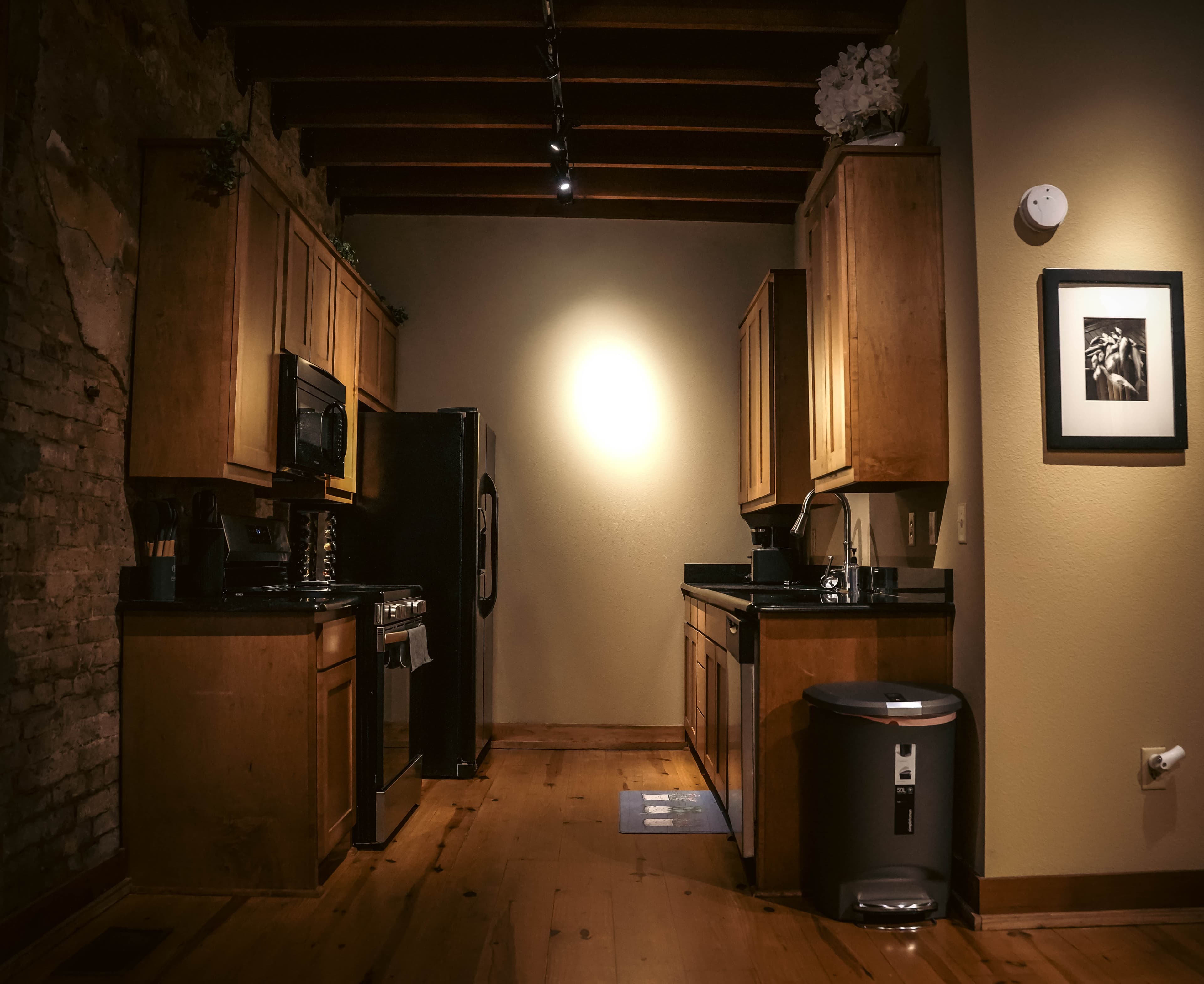 Narrow, dimly lit galley kitchen with warm wood cabinets, black appliances (including a refrigerator), exposed ceiling beams, and a bright spotlight on the far wall.