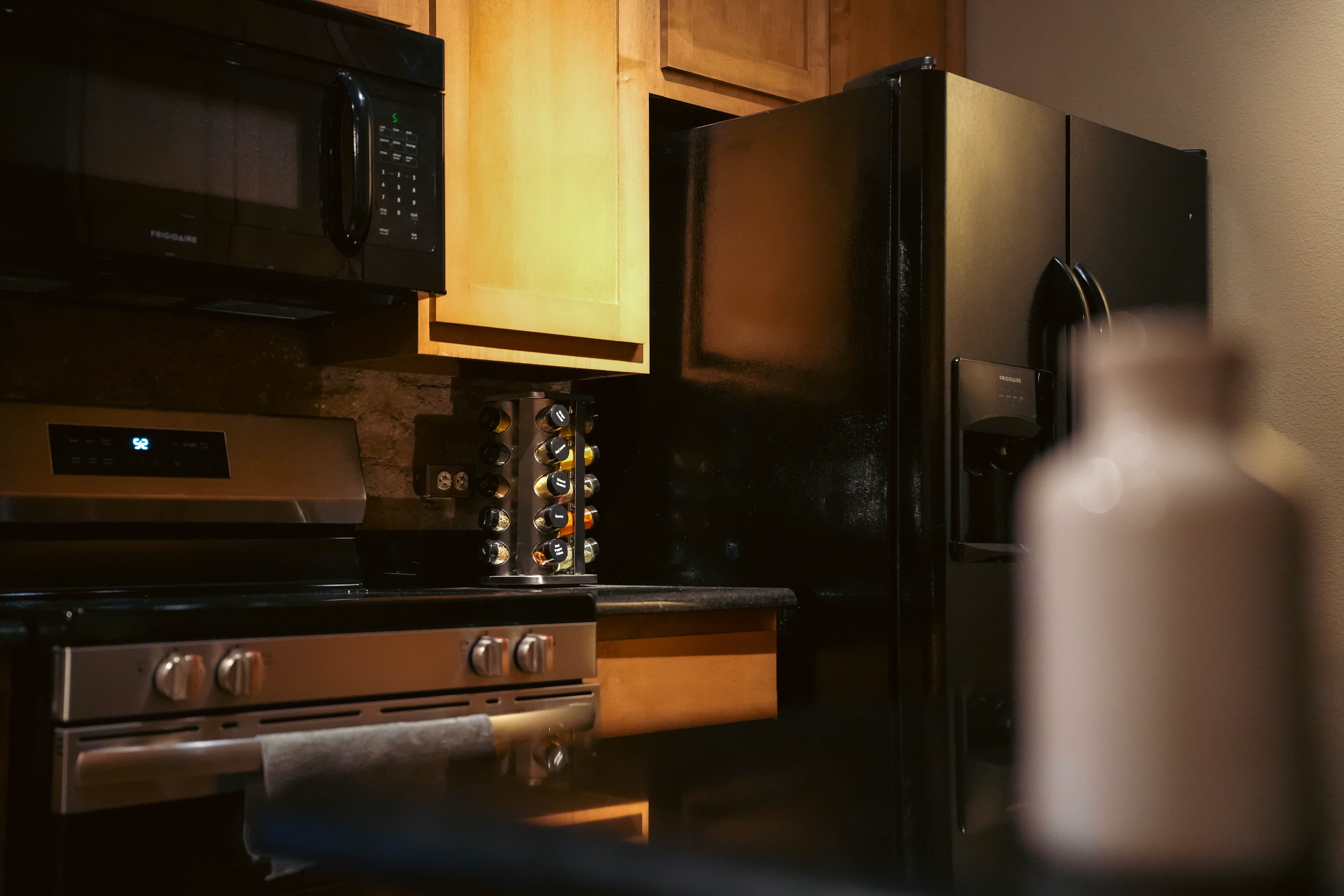Close-up of a kitchen corner with stainless steel and black appliances, including a range and microwave, light wood cabinets, and a black refrigerator.