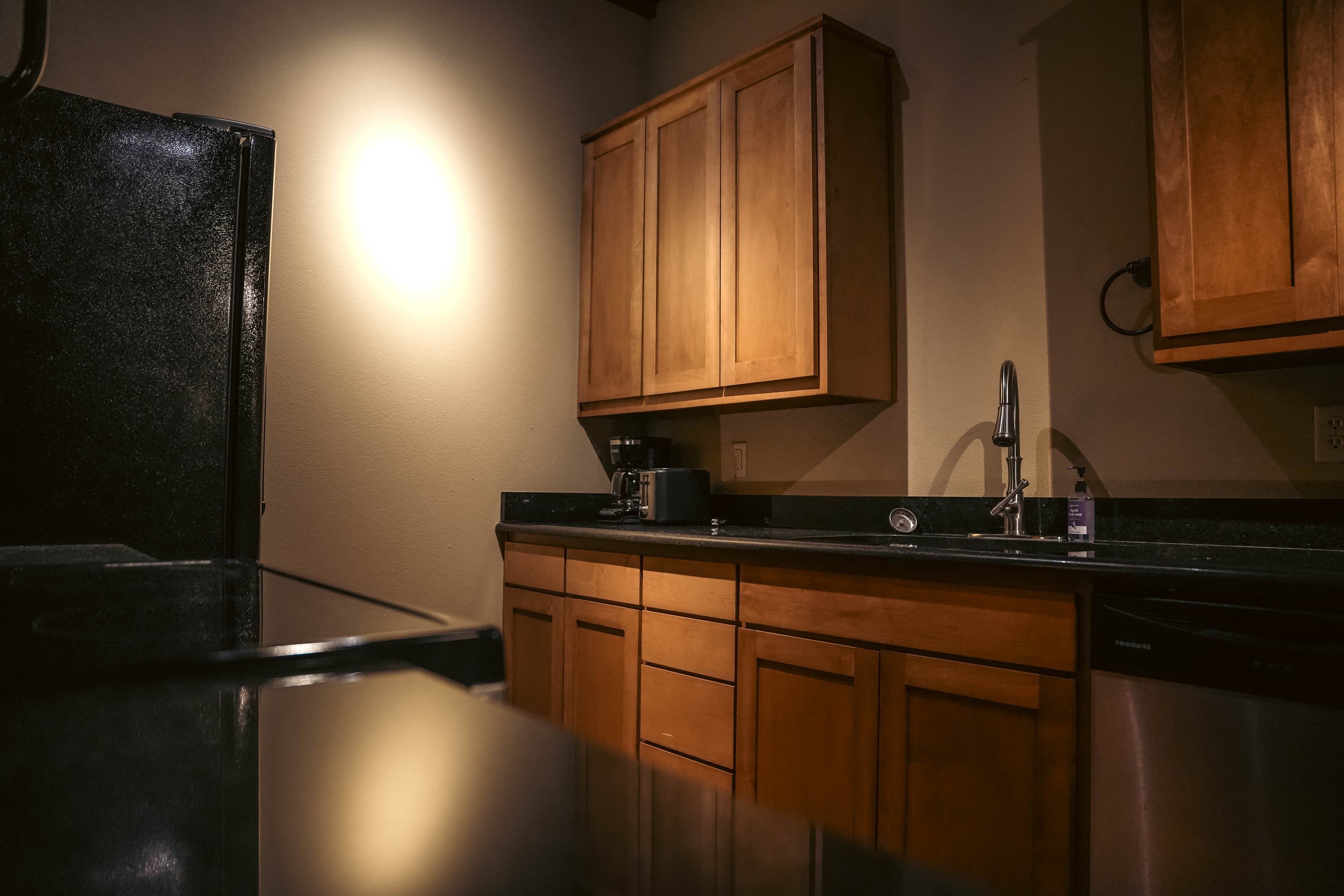 Cozy, dimly lit kitchen corner featuring wood cabinets, dark black countertops, a sink with a gooseneck faucet, and a black refrigerator visible on the left.