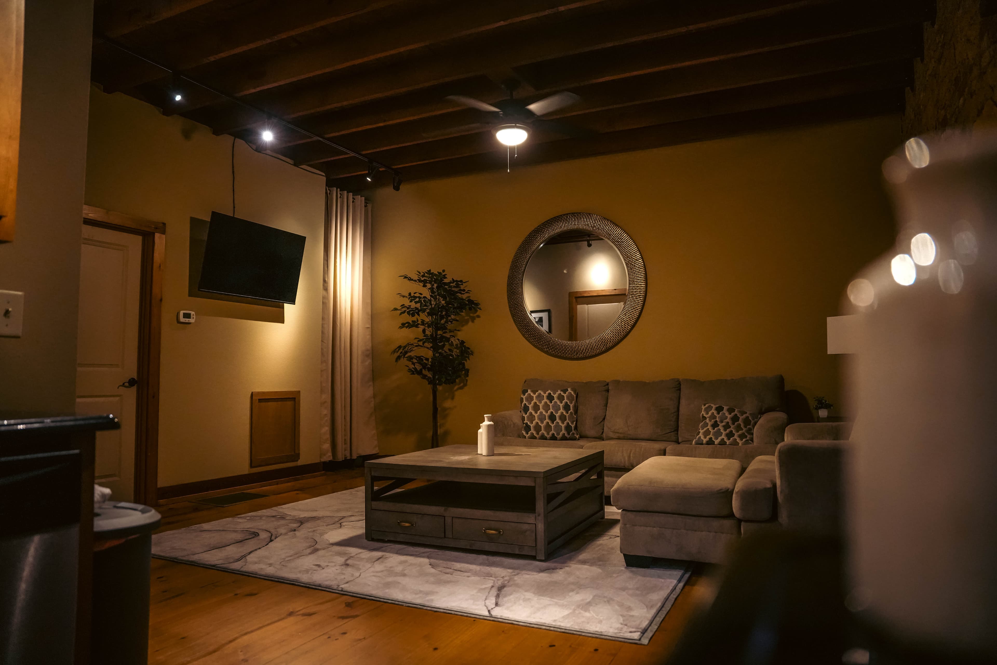 Cozy living room with a sectional sofa, a rustic wooden coffee table, a large round mirror, a wall-mounted TV, and exposed wood ceiling beams.