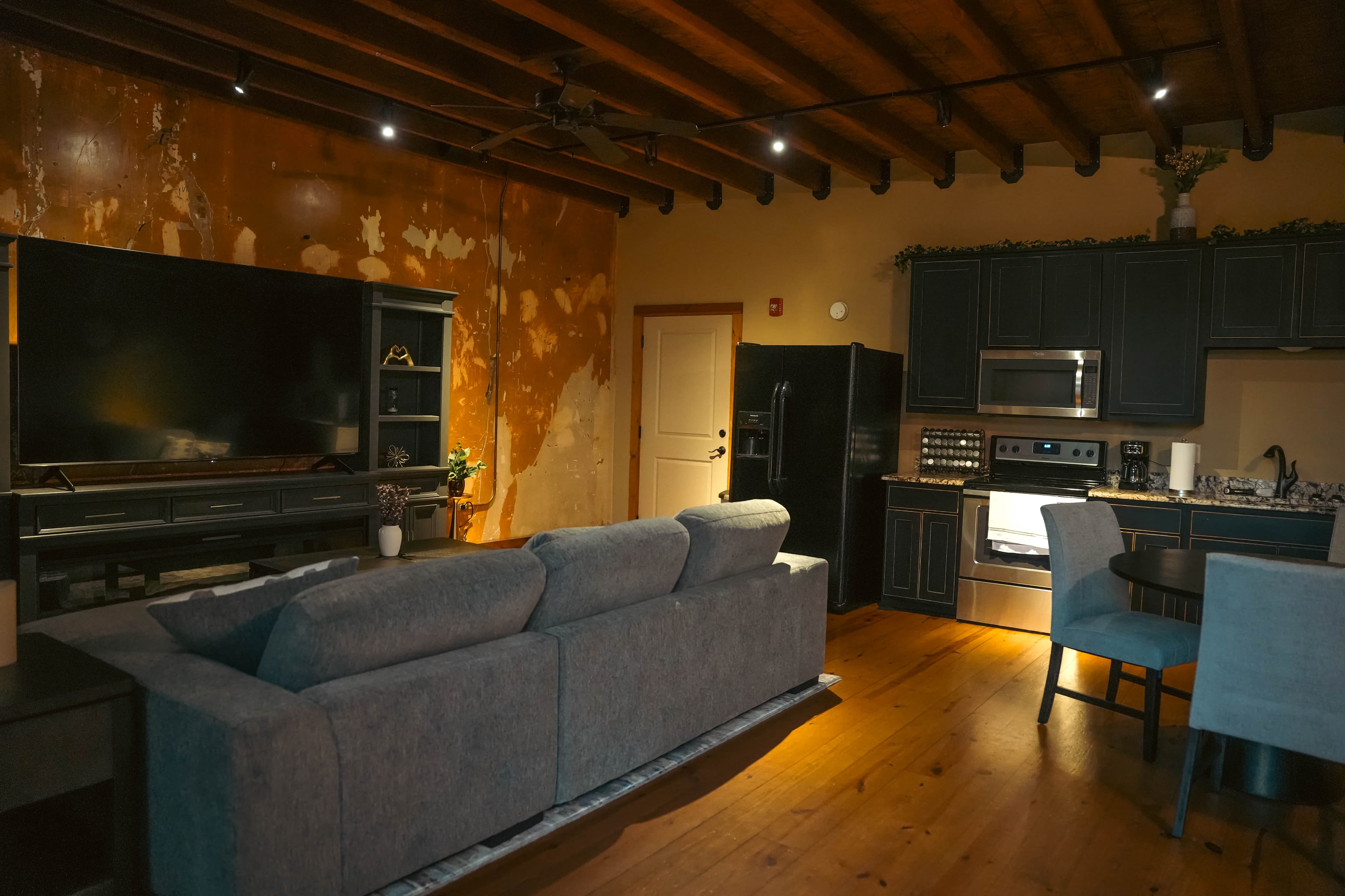 Open-concept apartment with a gray sofa, a large entertainment center and TV, dark kitchen cabinets, exposed wood ceiling beams, and distressed walls.