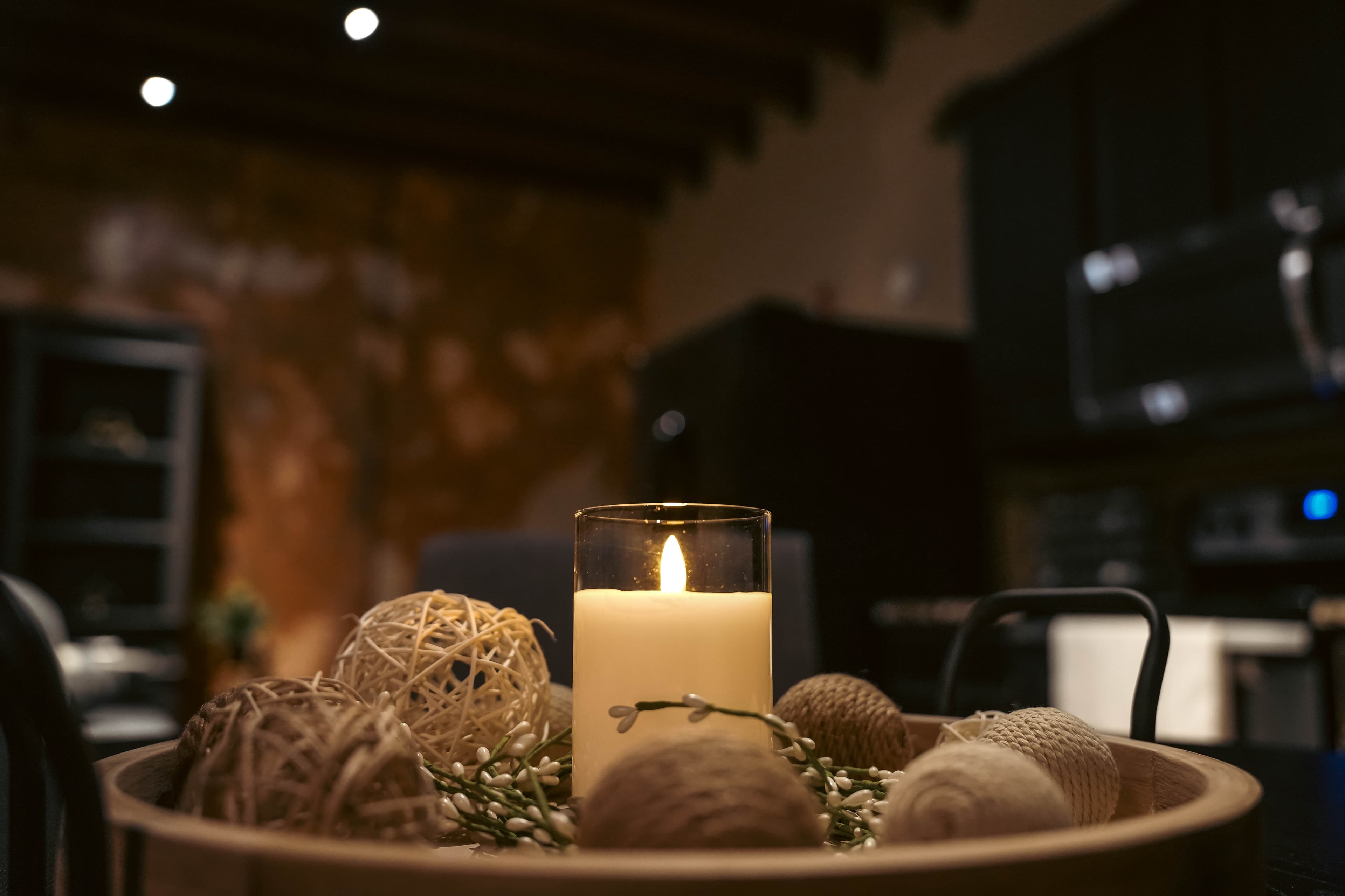 Close-up of a lit candle and rustic twine balls in a wooden tray, with a blurred dark kitchen and exposed wood ceiling beams in the background.