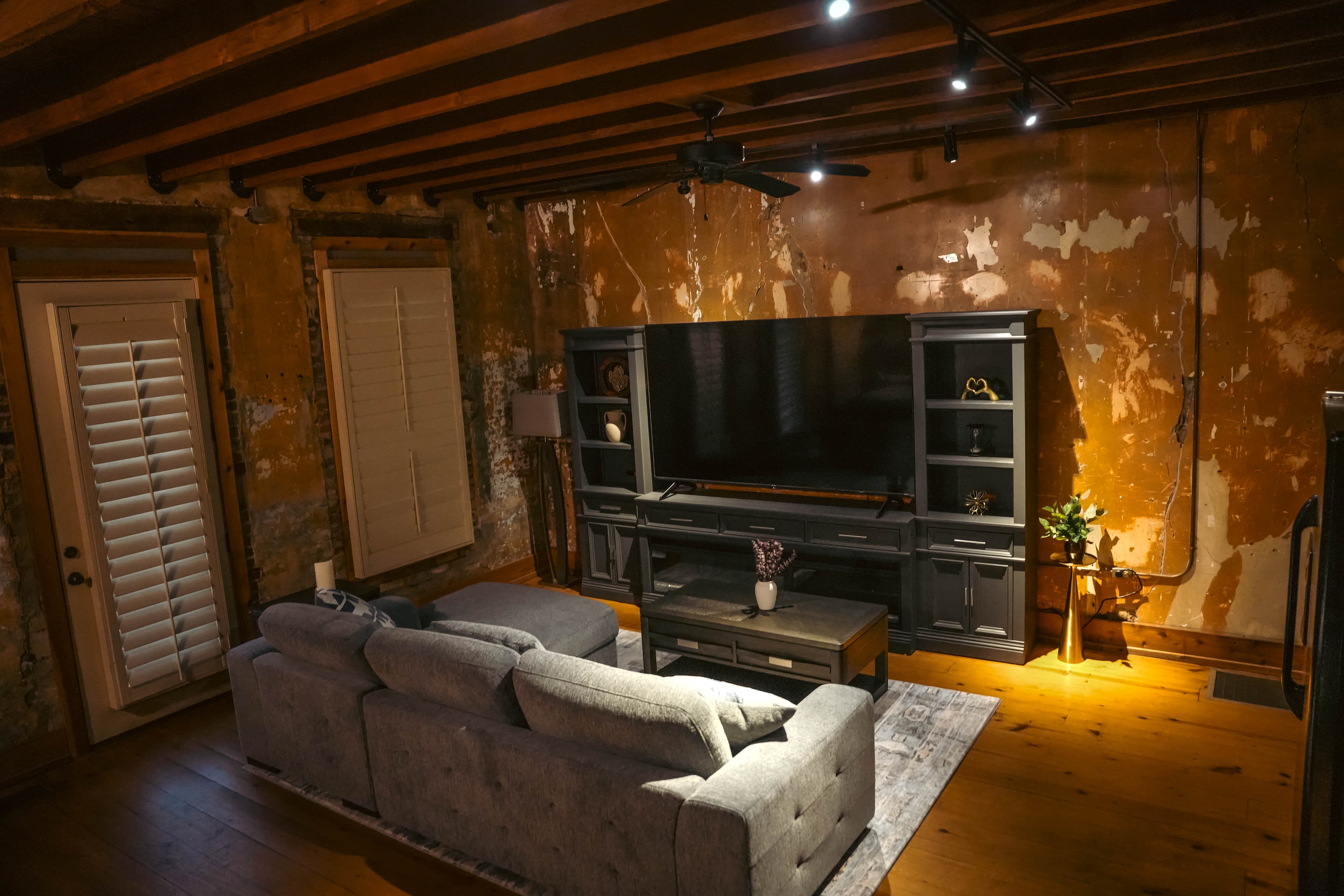 Rustic industrial living room featuring a gray sofa, a dark media console with a large TV, distressed walls, exposed ceiling beams, and wood flooring.