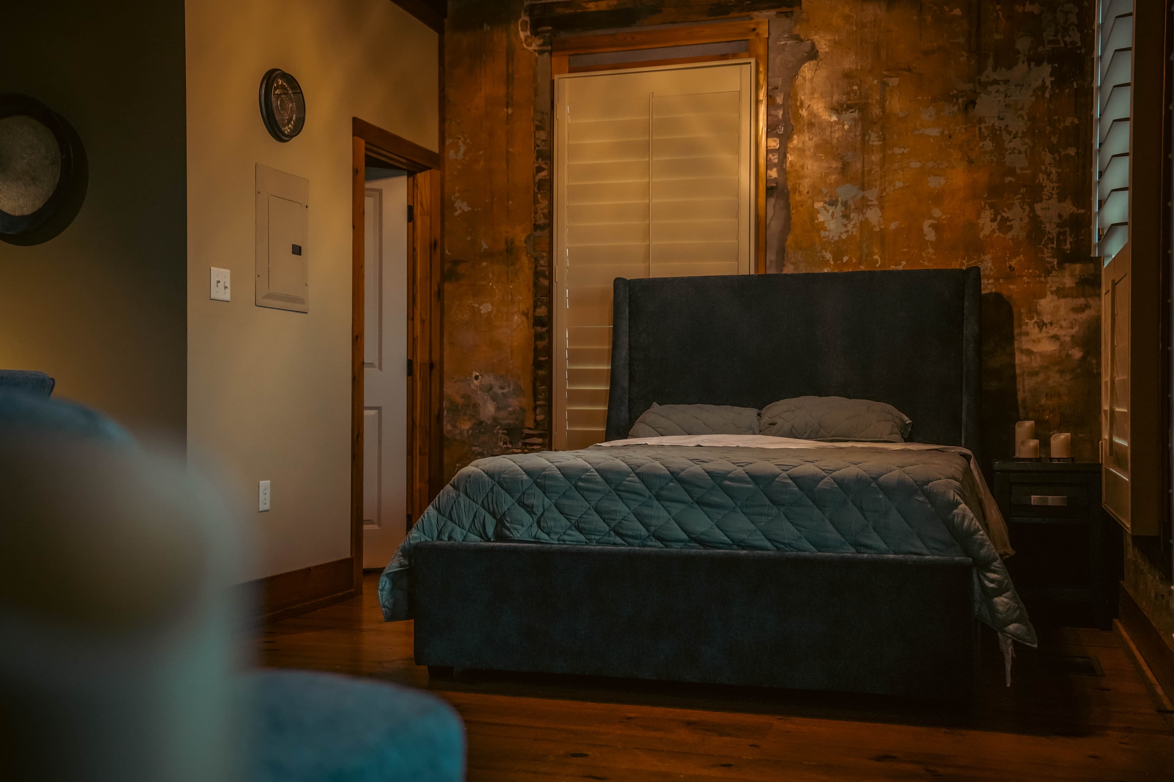 Industrial-style bedroom area with a dark upholstered bed, distressed brick walls, wood floors, and a glimpse of a nearby doorway.