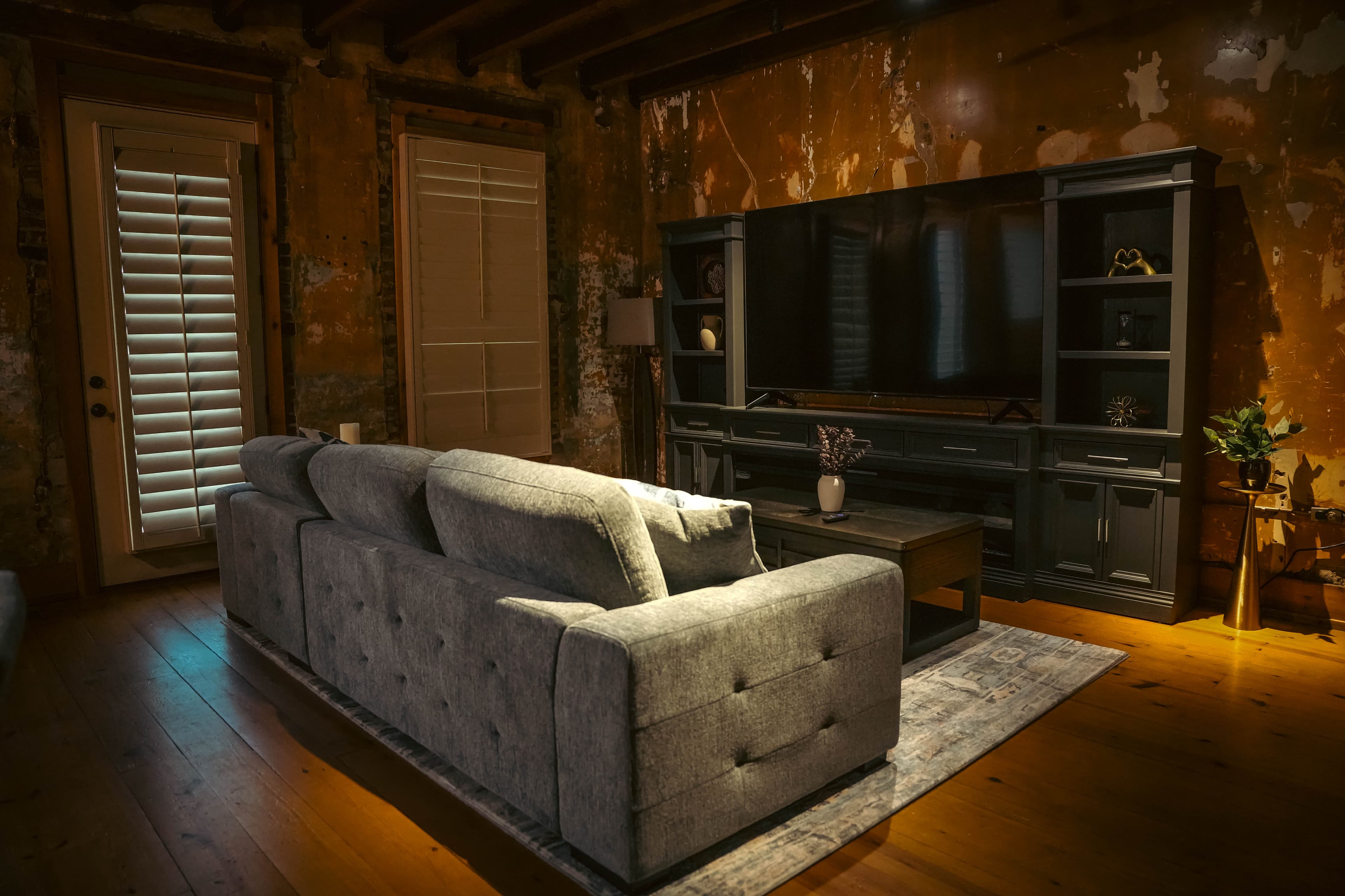 Rustic living room with a gray velvet sofa, a dark media console and large TV, distressed brick walls, and exposed wood ceiling beams.