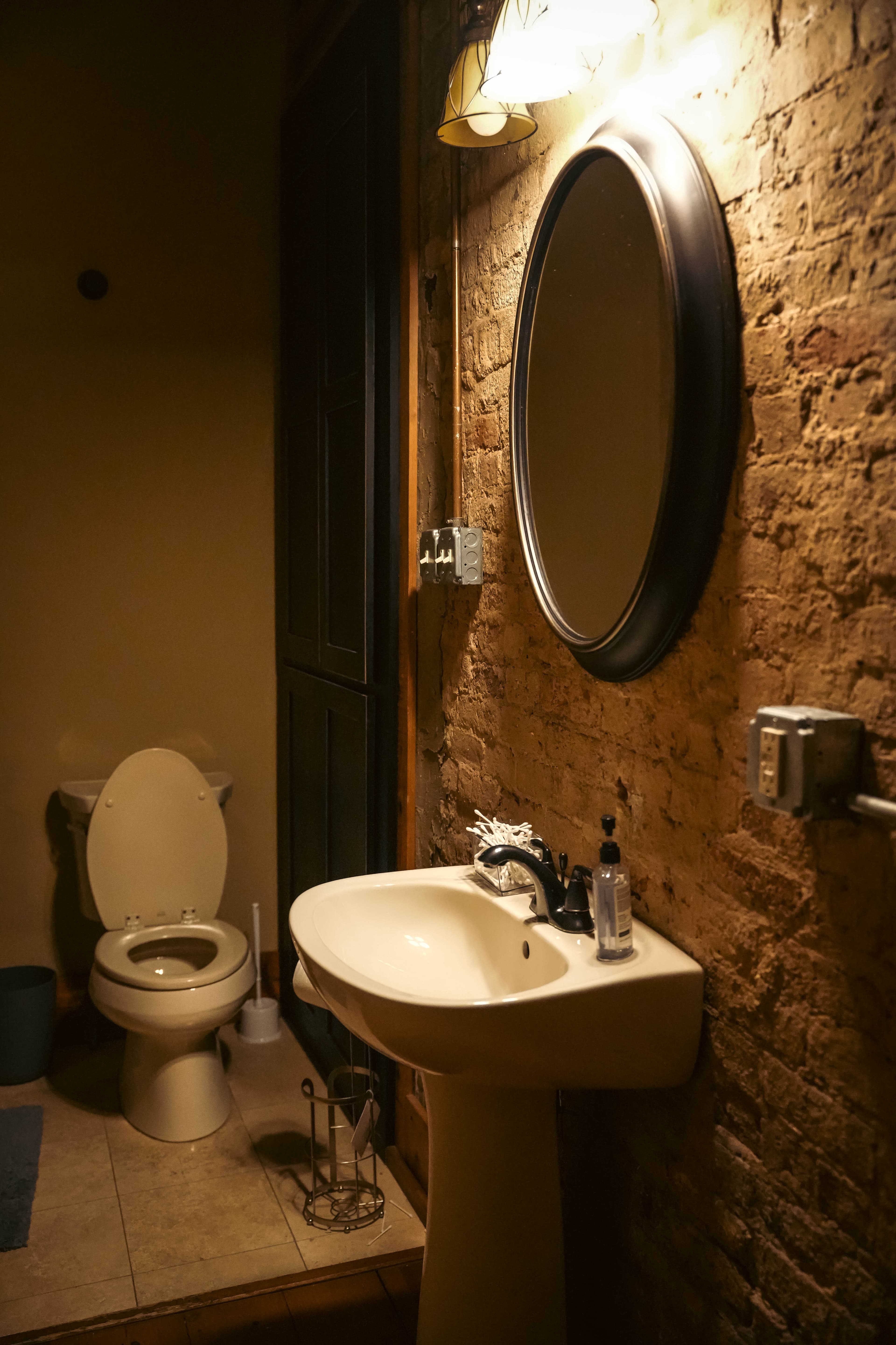 Rustic bathroom featuring a white pedestal sink, an oval mirror, a light fixture above it, and an exposed brick wall next to the toilet.