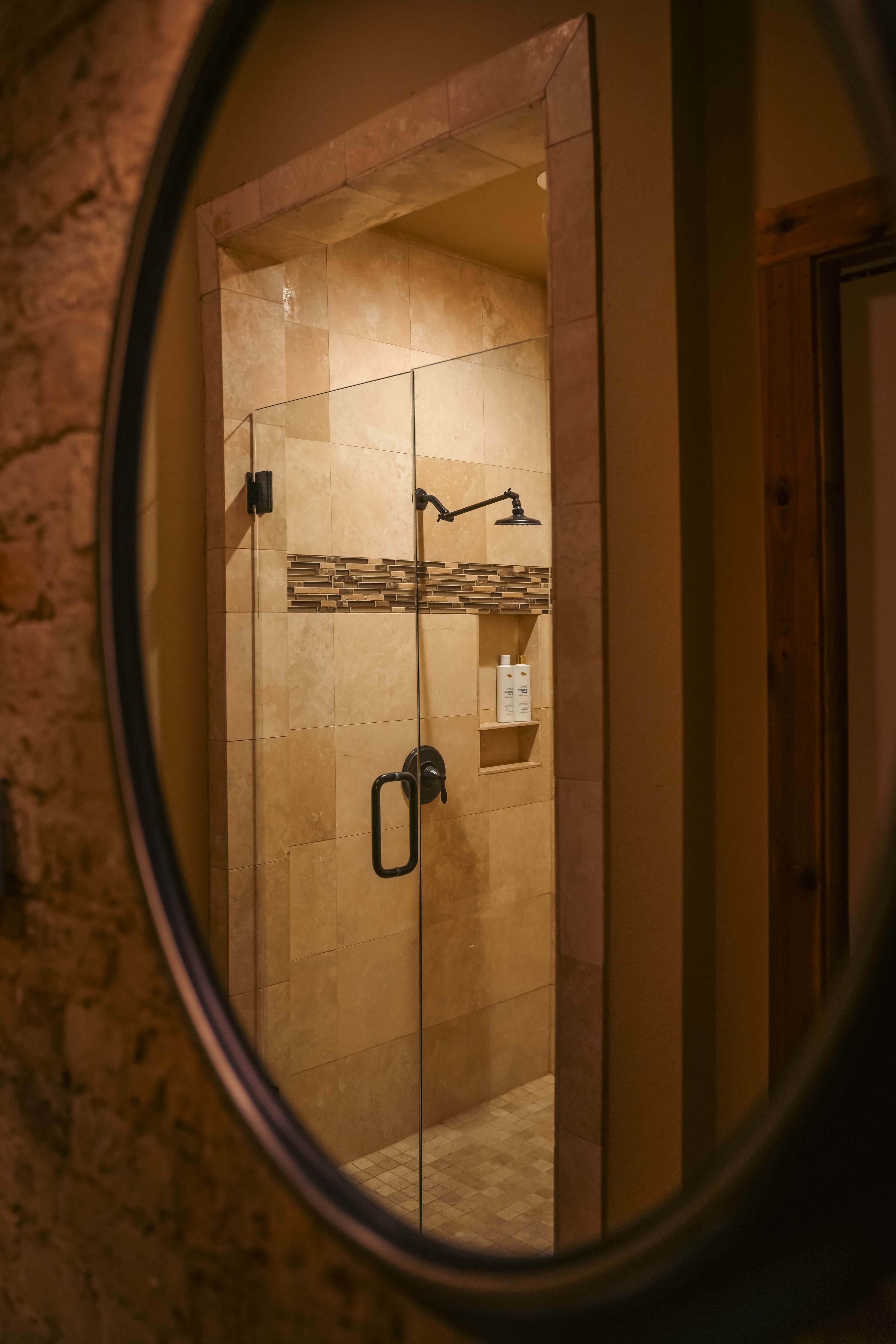 Bathroom image reflecting a modern, tiled walk-in shower with a glass door and decorative tile strip, viewed through a large, oval mirror.