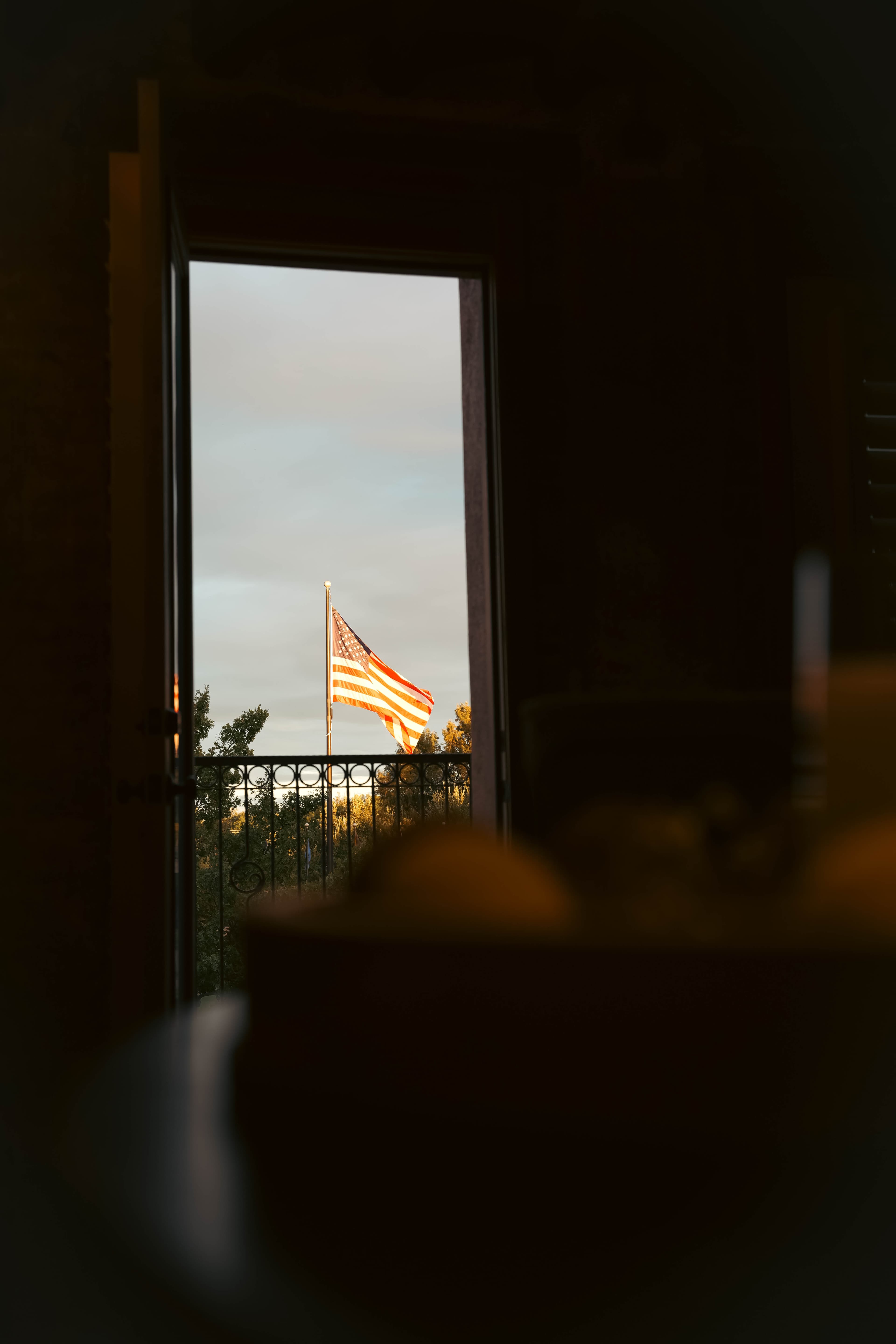 View of an American flag waving on a pole, framed by an open balcony door, with a dark interior foreground and sunset light hitting the flag.