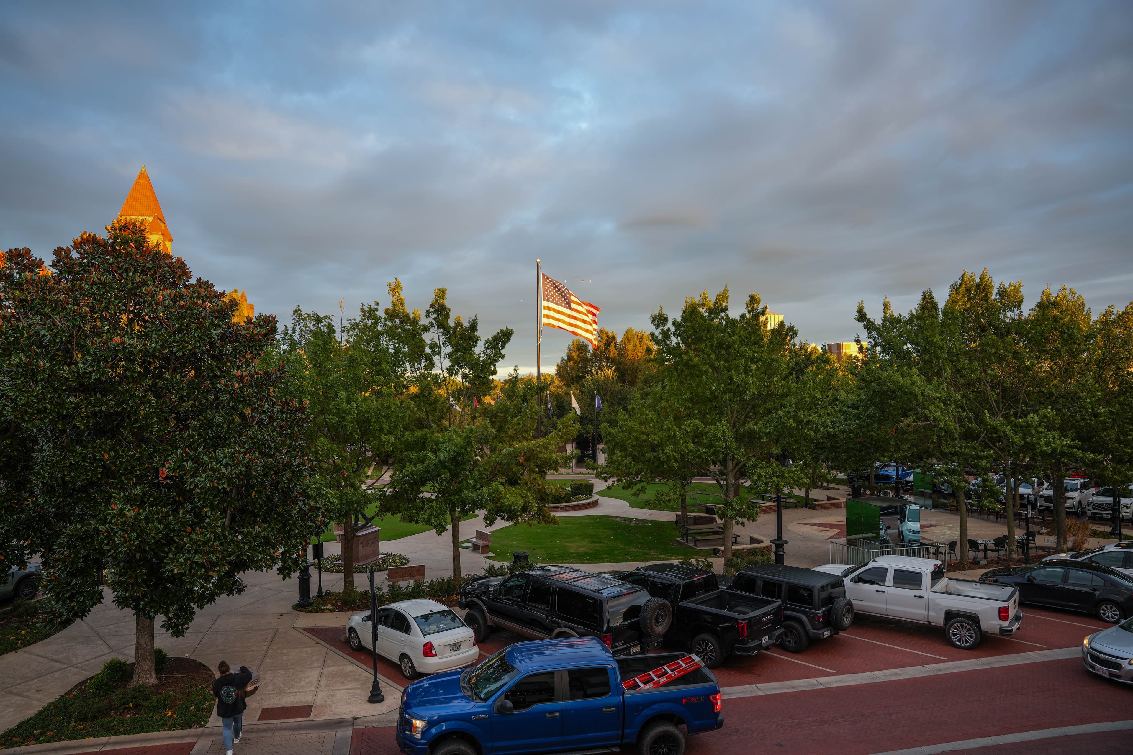 Aerial view of a town square with green trees, brick-paved streets, parked cars, and a flagpole with a slightly visible American flag under a cloudy sky.