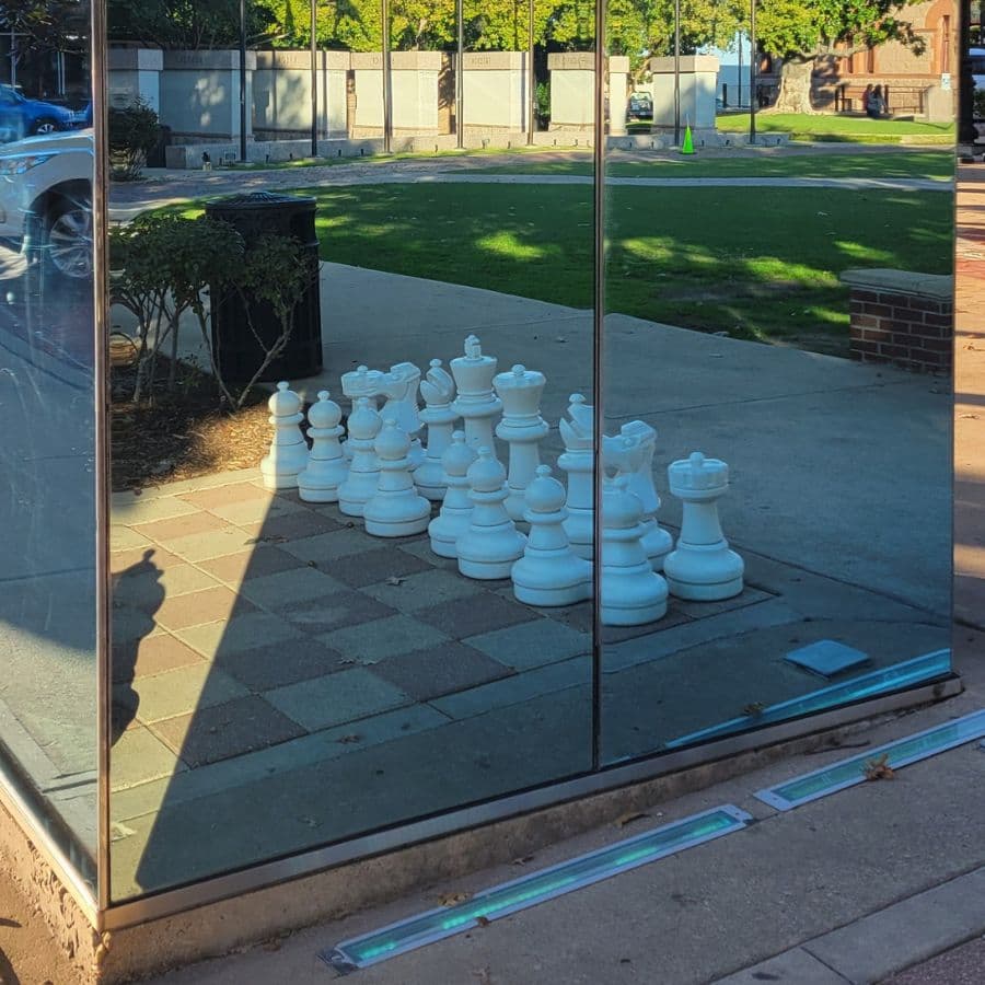 A reflection of large white chess pieces displayed on a sidewalk.