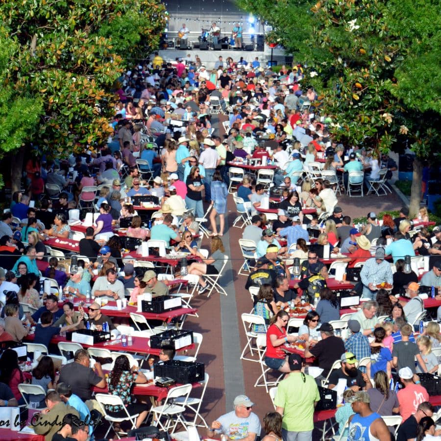 A crowded outdoor dining event with numerous tables filled with people enjoying food and live music.