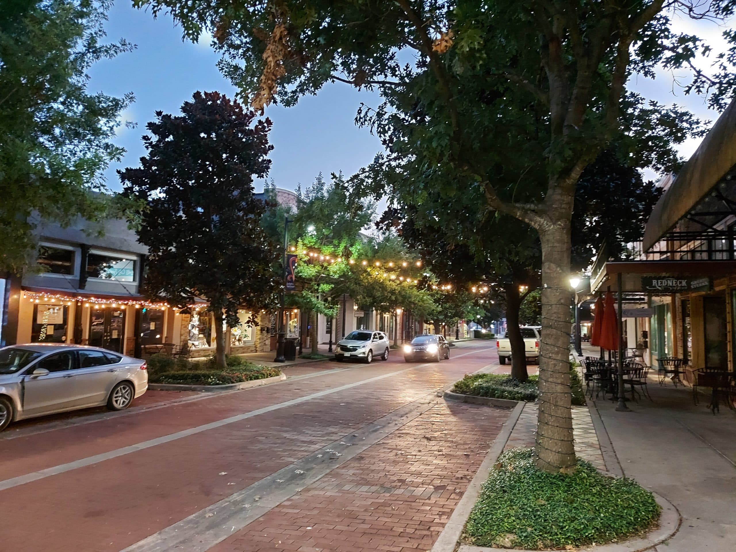 A charming street lit by string lights, lined with trees and shops in the early evening.