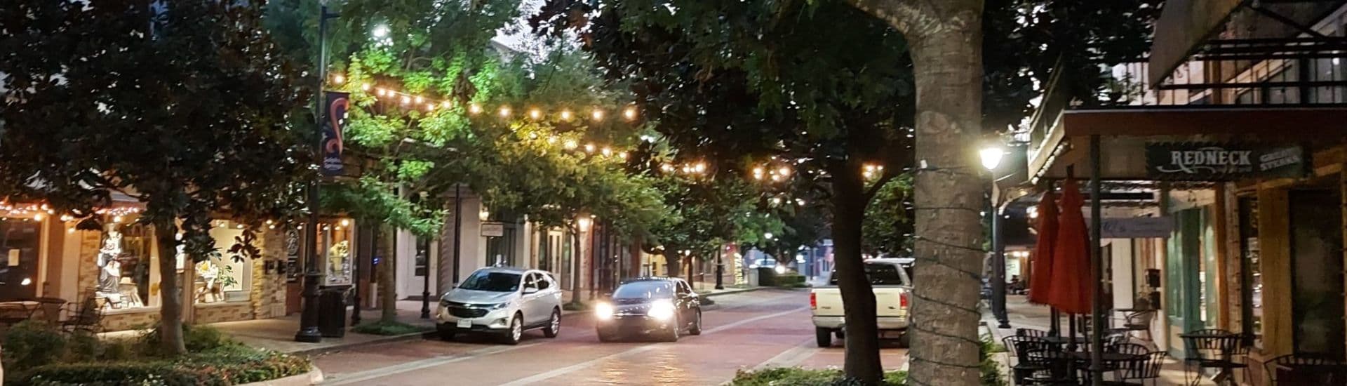 A serene nighttime street lined with trees and string lights, featuring parked cars and shops.