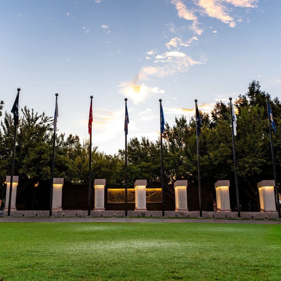 A row of flags stands in front of a monument on a grassy area under a colorful sky.