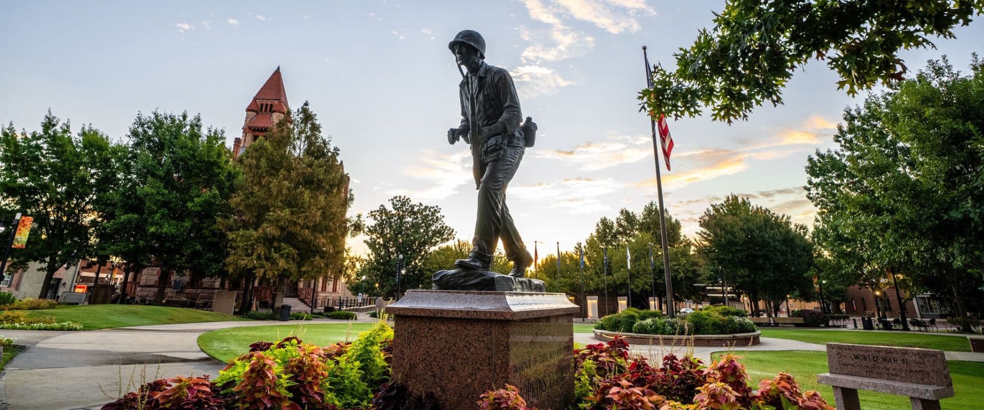 A soldier statue stands in a park surrounded by greenery and a sunset backdrop.