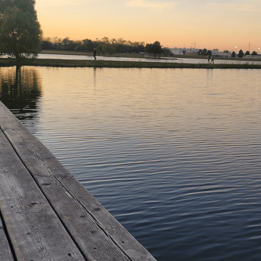 A peaceful lakeside scene at sunset with wooden planks in the foreground and silhouettes of people walking along the shore.