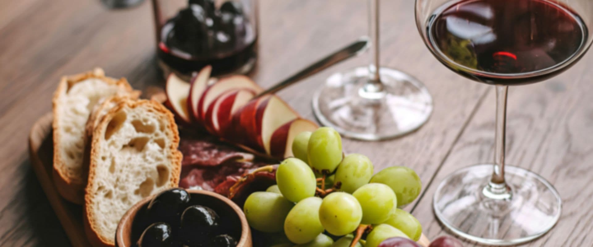 A wooden platter with bread, assorted grapes, olives, and slices of apple, accompanied by two glasses of red wine.