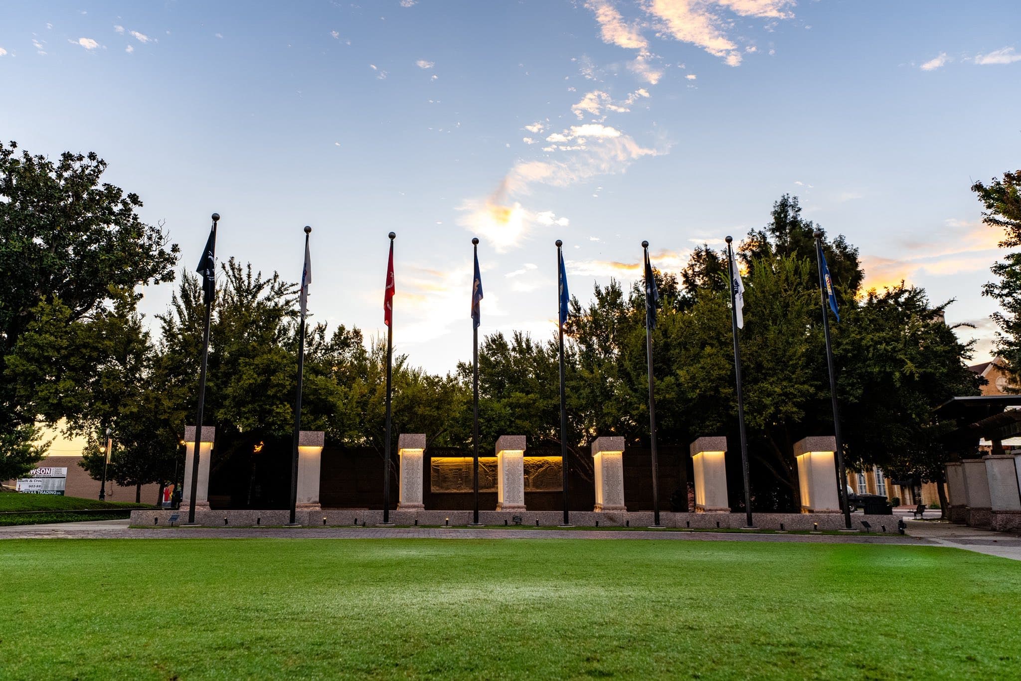 A row of flags stands in front of a memorial surrounded by green grass under a sunset sky.