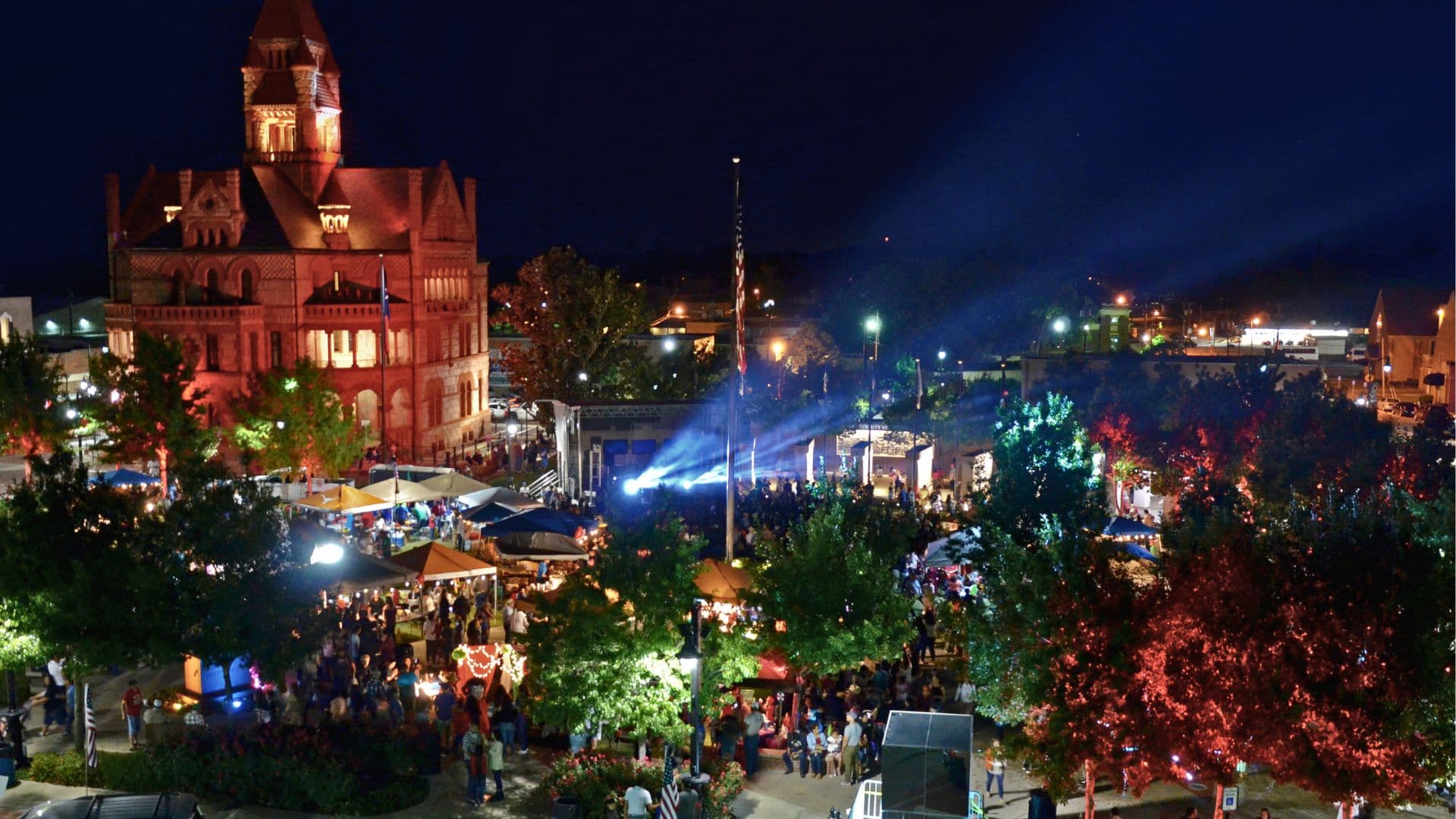 A vibrant night market crowd gathers near a historic building, illuminated by colorful lights and search beams.