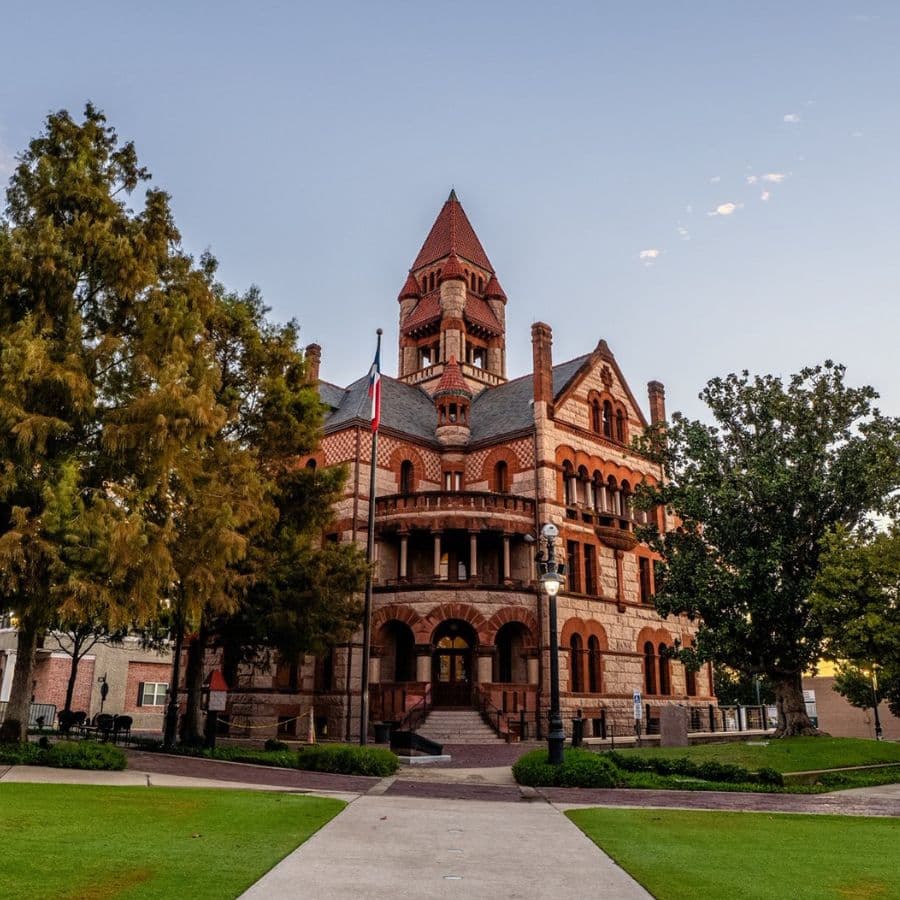 Historic red-brick building with a tall turret surrounded by trees and a manicured lawn.