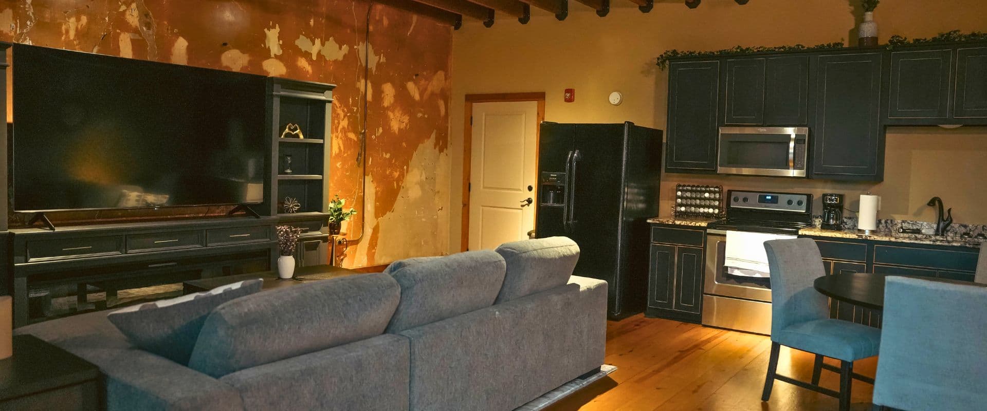 A cozy living room featuring a gray sofa, large TV, and a modern kitchen with dark cabinetry.